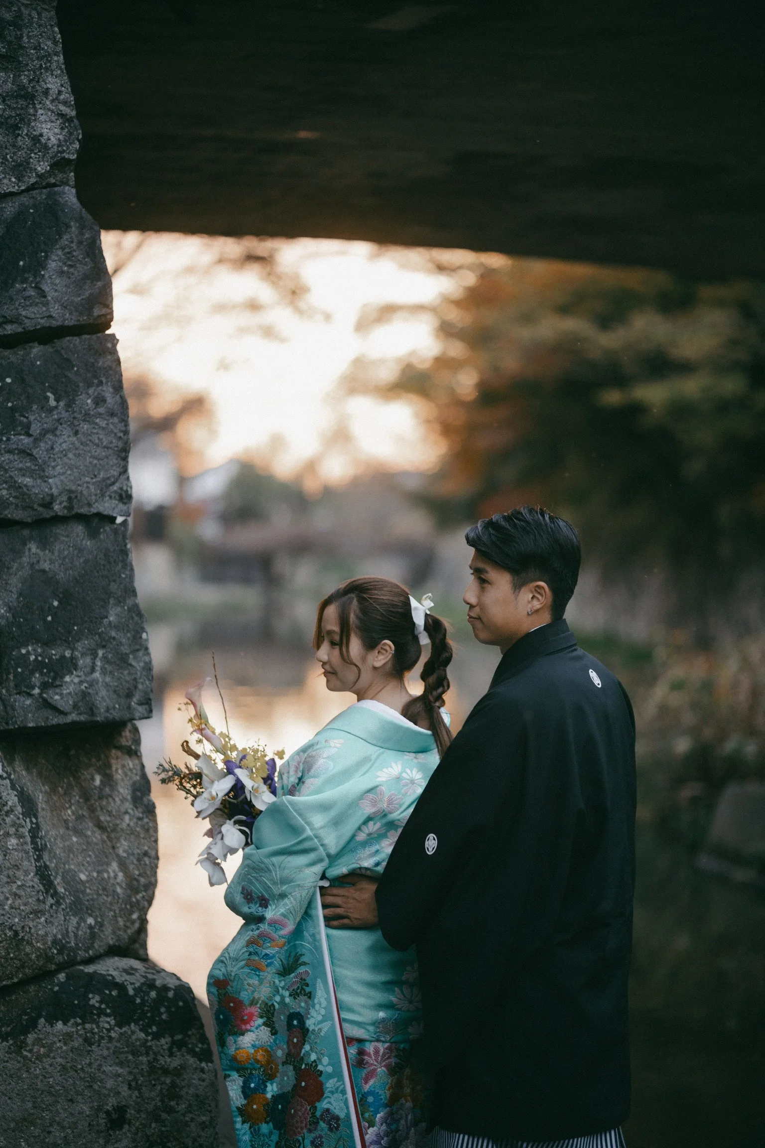 A couple dressed in traditional Japanese wedding attire standing under a bridge near a river at sunset.