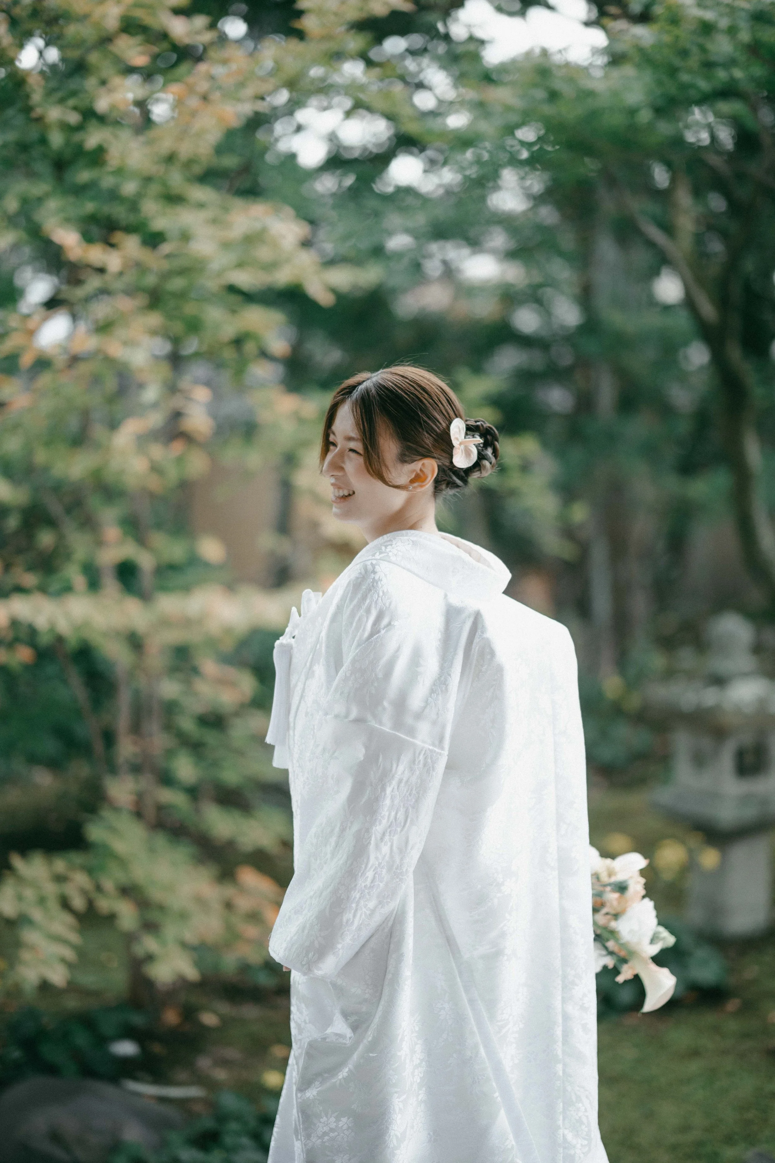A woman in a white traditional dress standing outdoors, smiling, with greenery in the background.
