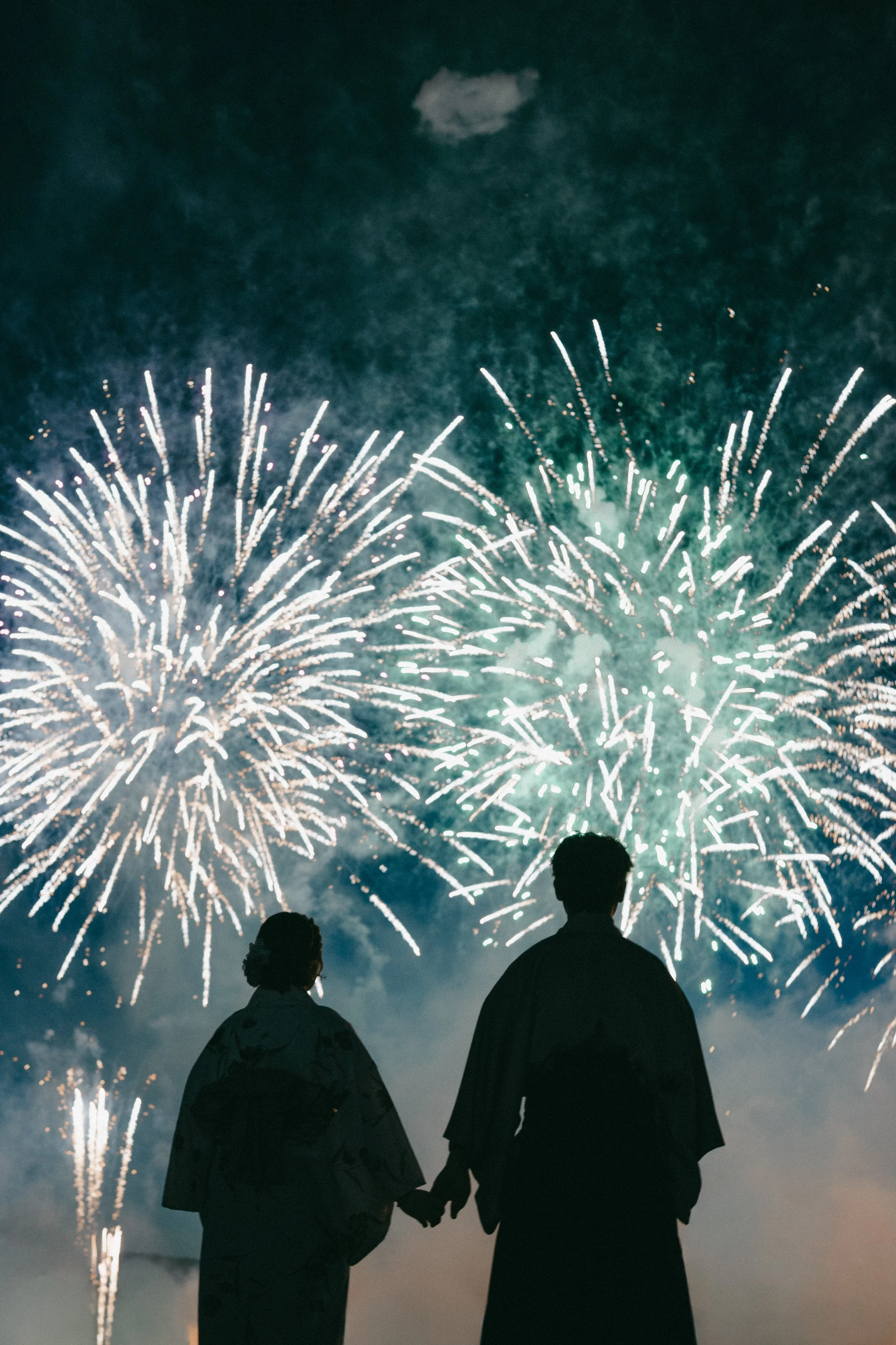 Silhouettes of a couple holding hands and watching fireworks in the night sky.