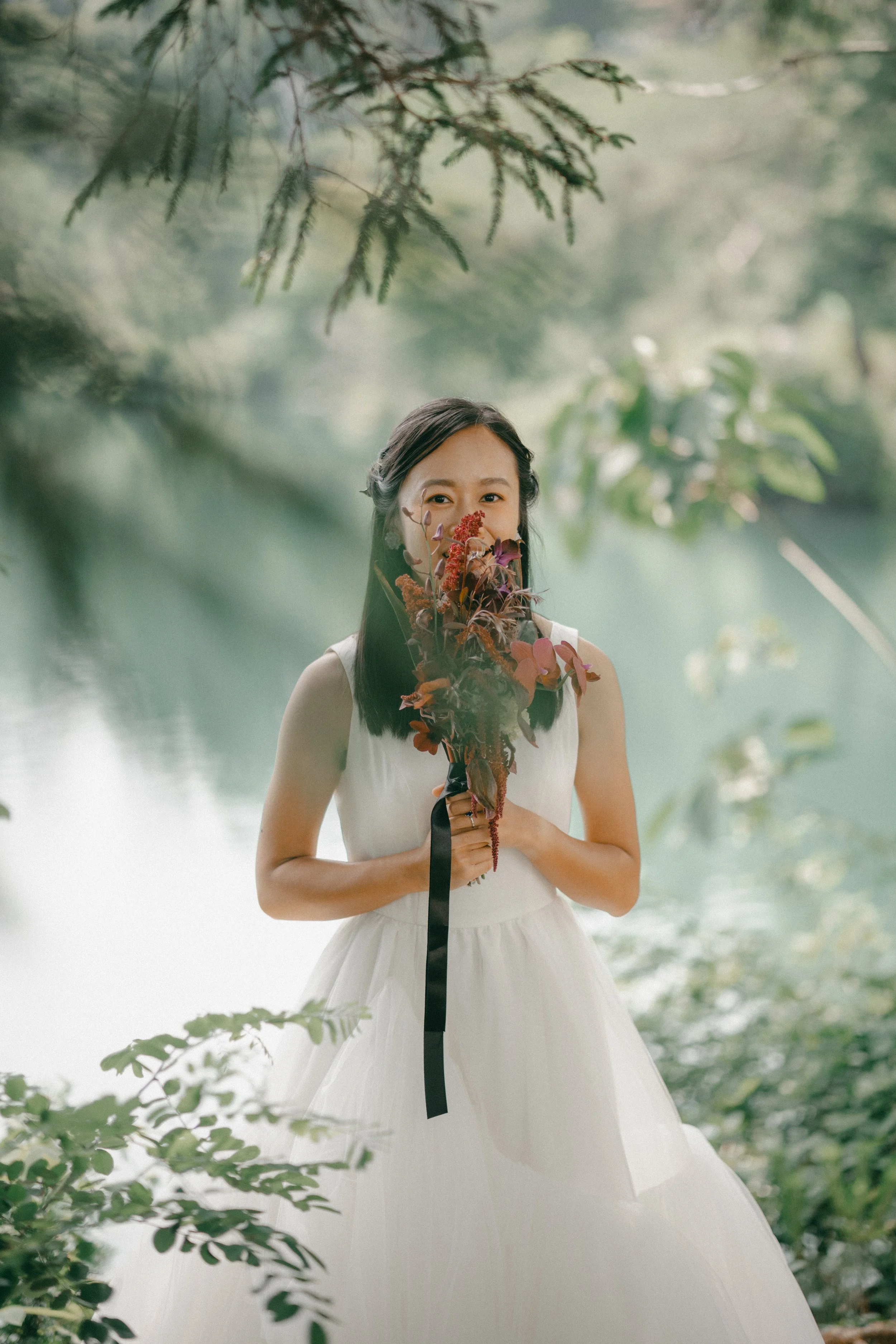 A woman in a white dress holding a bouquet of flowers in front of her face, standing outdoors near a body of water, surrounded by green foliage and trees.