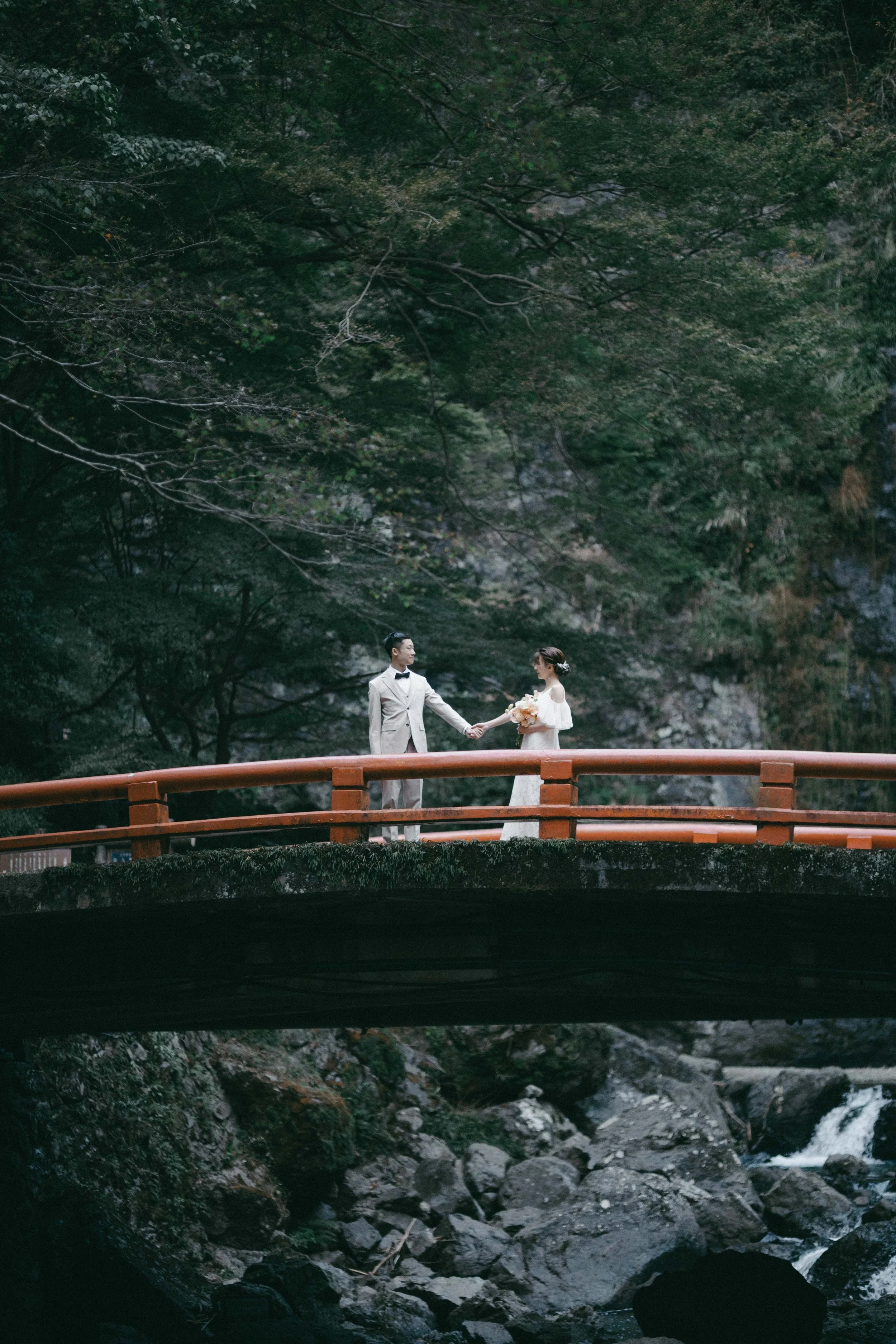 A bride and groom holding hands on a red arched bridge over a rocky creek, surrounded by dense green trees.
