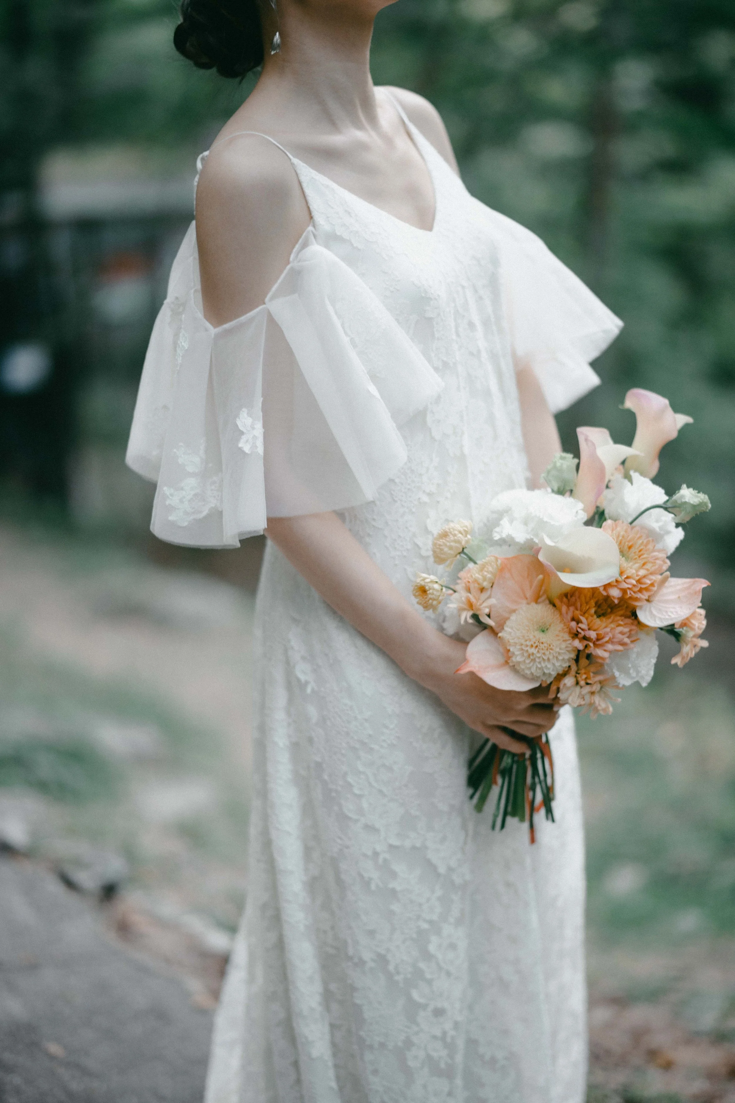A woman in a white lace dress holding a bouquet of peach and white flowers outdoors.