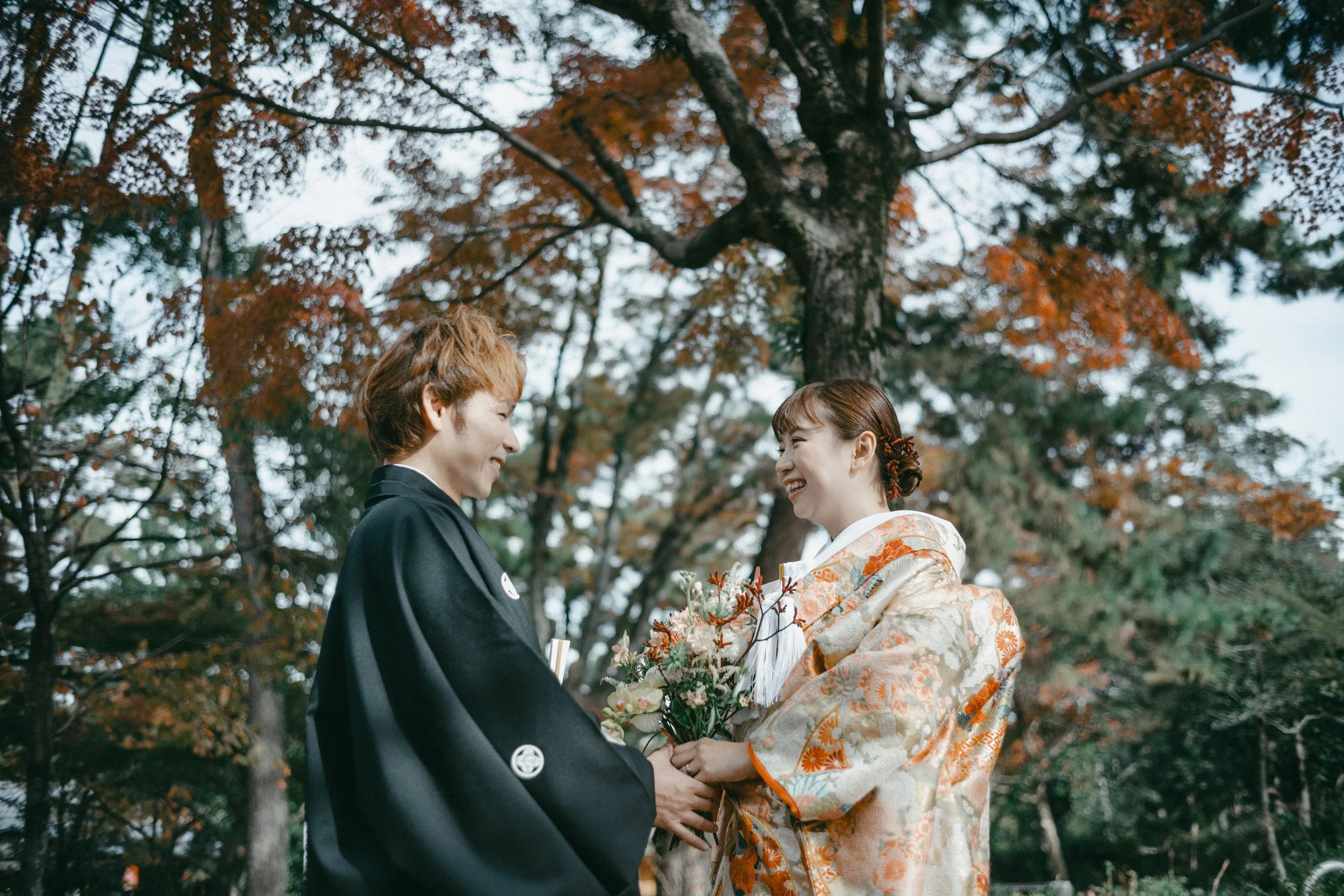 A couple in traditional Japanese wedding attire sharing a joyful moment outdoors among autumn trees, with the bride holding a bouquet of flowers.
