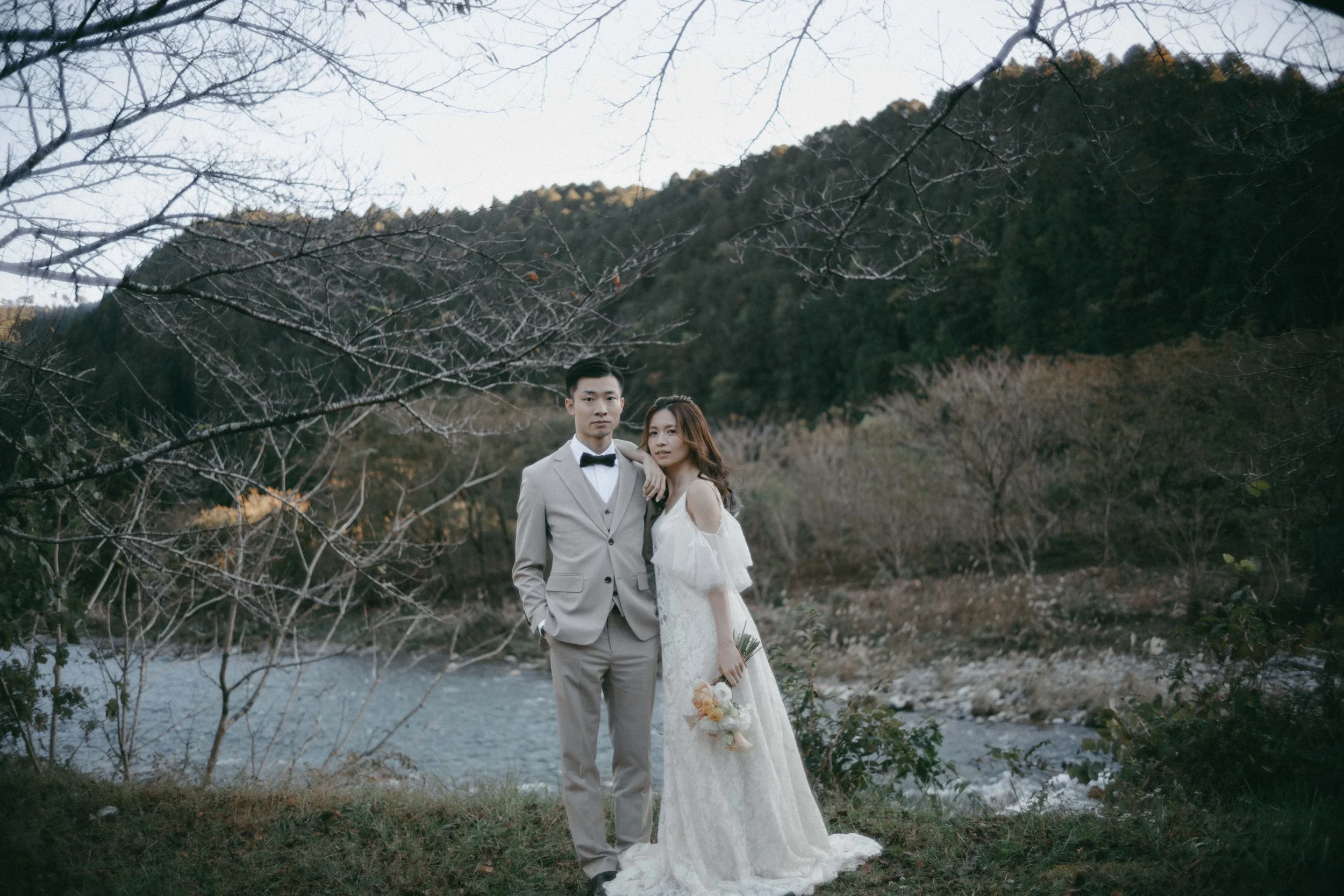 A couple dressed in wedding attire standing outdoors near a river with a mountain and trees in the background.