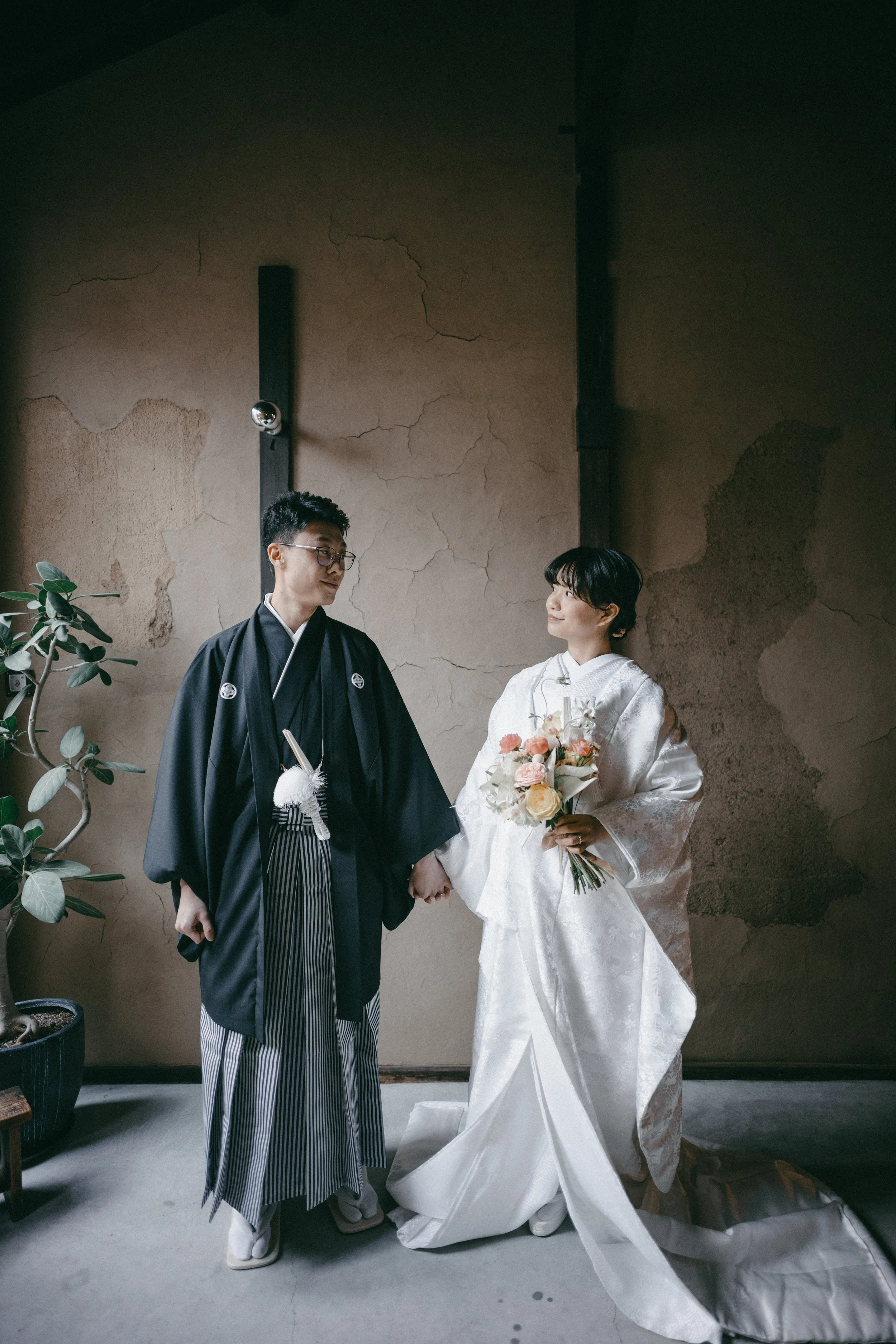 A couple dressed in traditional Japanese wedding attire holding hands, the bride holding a bouquet of flowers, standing indoors against a rustic wall with a potted plant nearby.