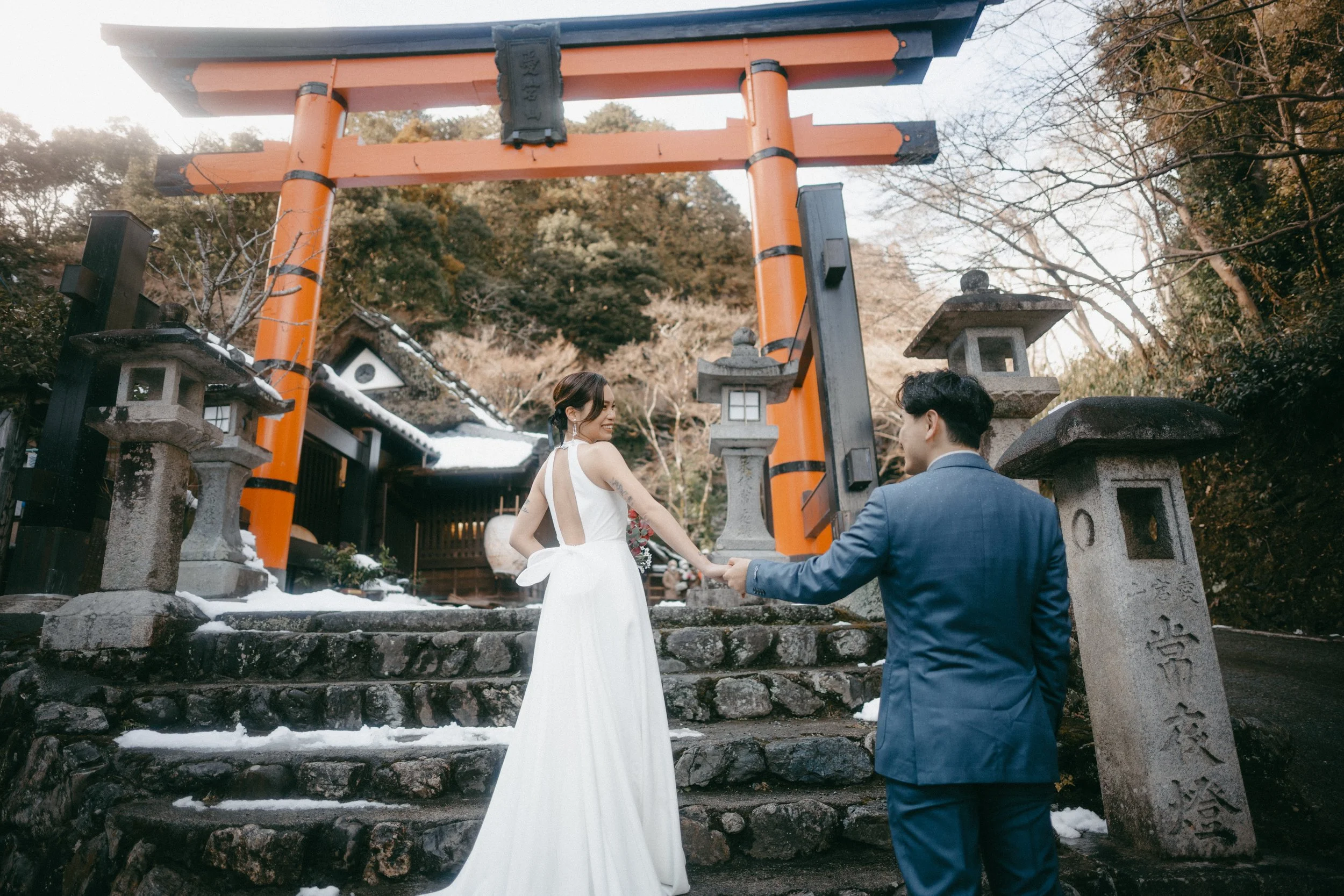 A couple in wedding attire holding hands on stone steps in front of a traditional Japanese torii gate and lanterns, with trees and a snow-covered ground in the background.