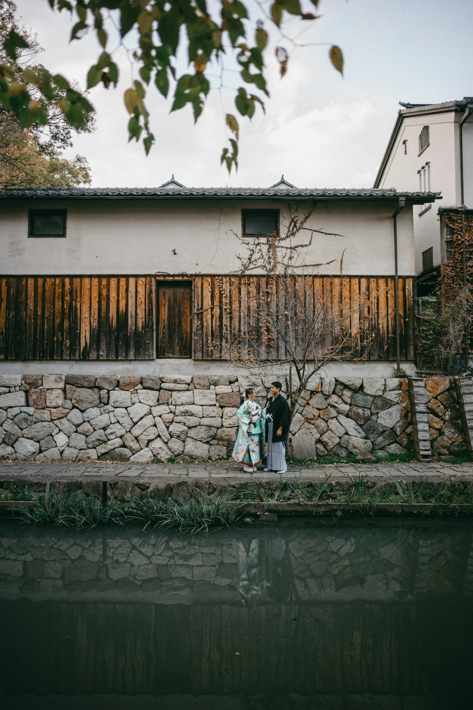 A Japanese couple in traditional clothing standing by a small tree near a stone wall and a waterway, with a building featuring a wooden fence in the background.