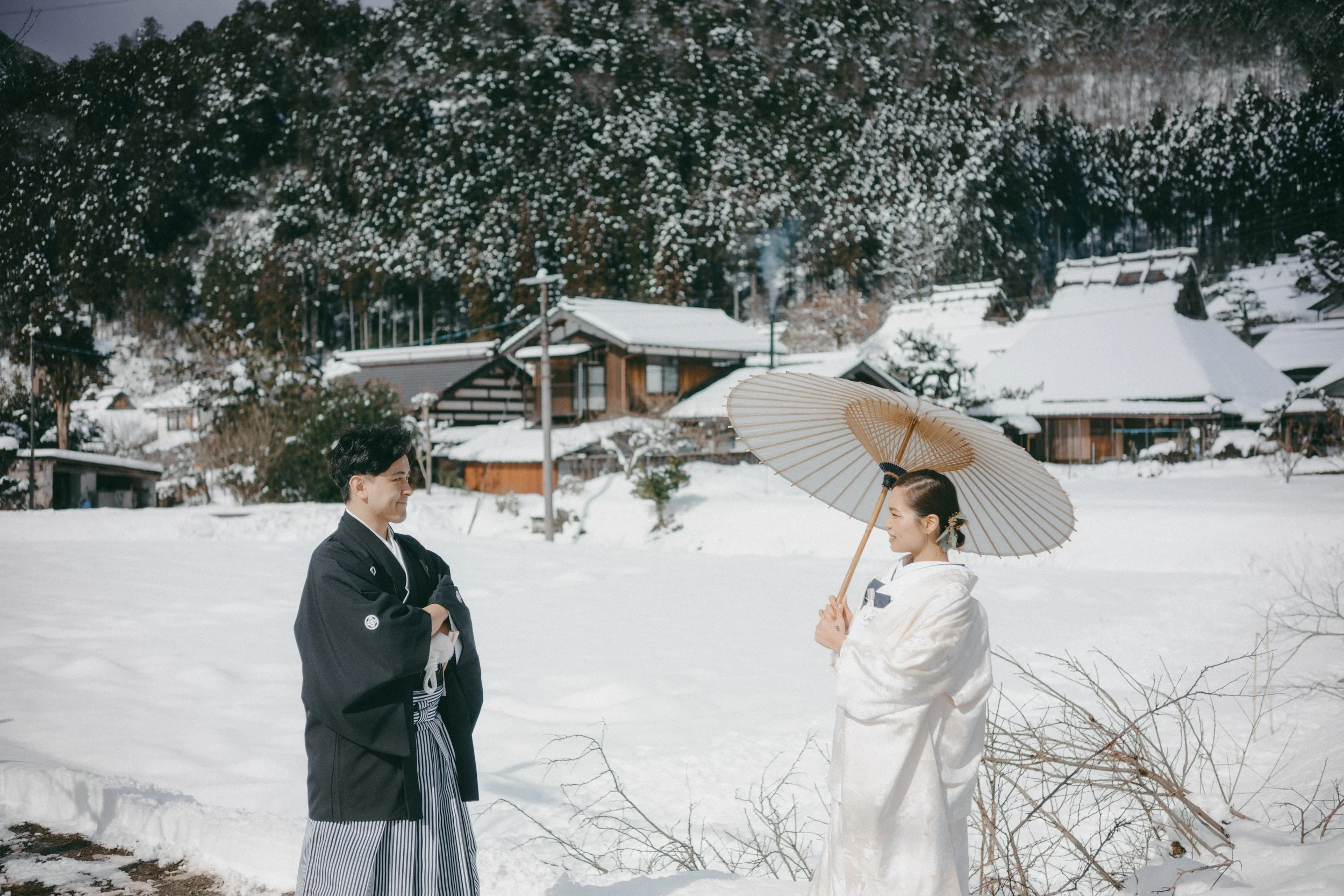 A couple in traditional Japanese wedding attire standing in a snowy landscape, facing each other. The woman holds a parasol over her head, and the man has his arms crossed, smiling. Traditional Japanese houses and snow-covered trees are in the backgr