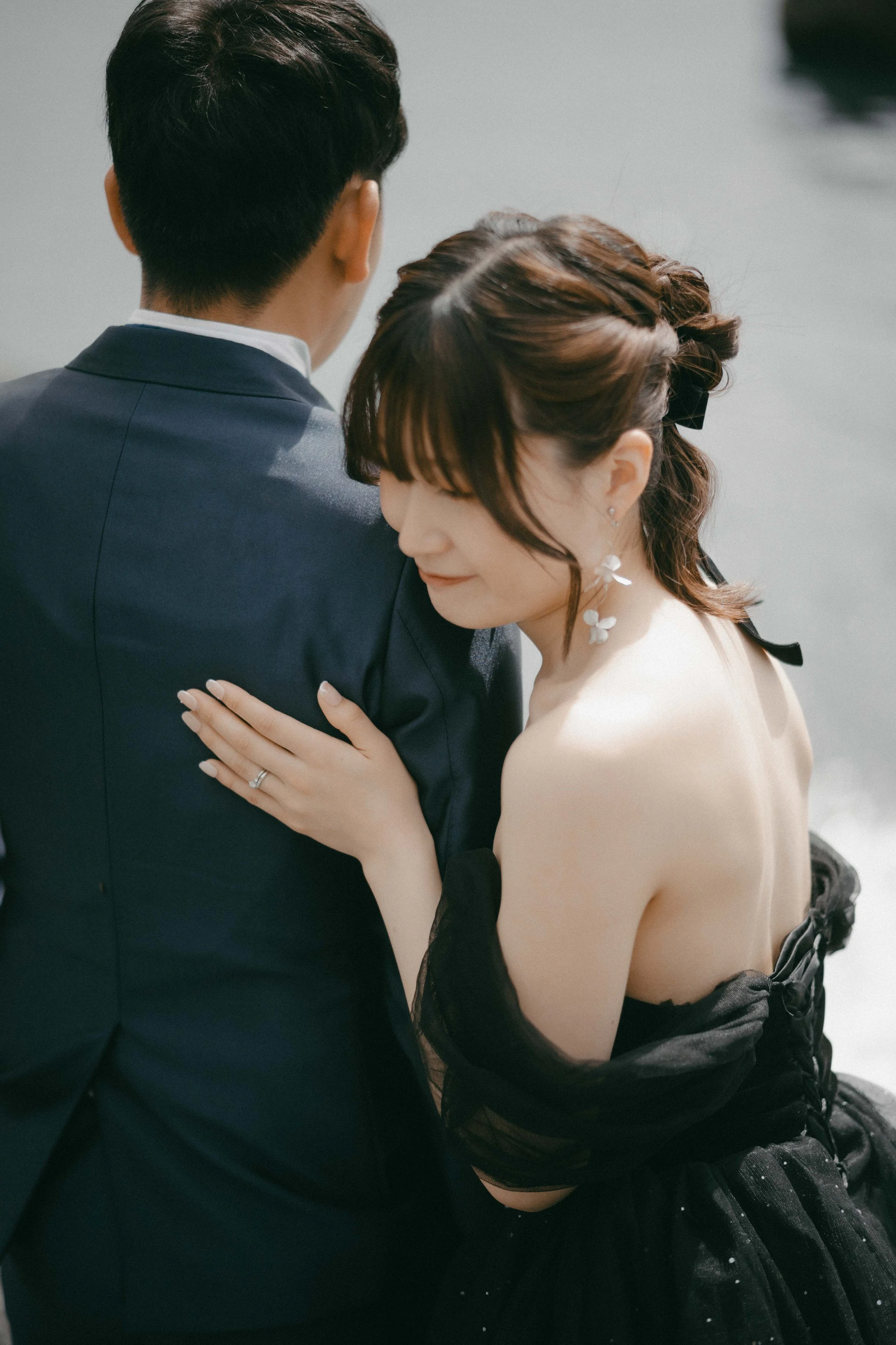 A woman with floral earrings and dark hair styled in waves, wearing a black off-shoulder dress, hugs a man in a navy suit from the back near the water.
