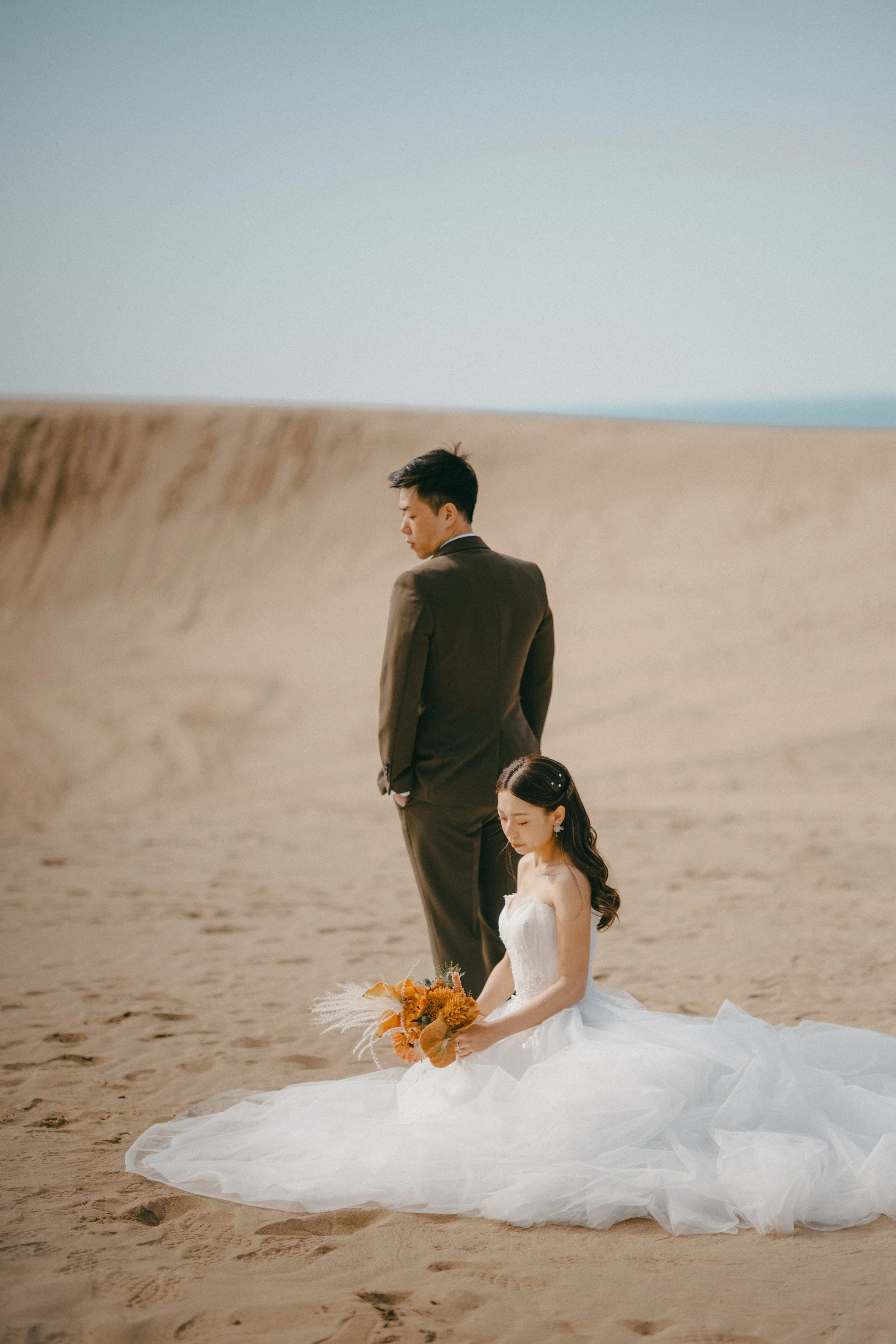 A bride in a white wedding dress sitting on the sand holding a bouquet, with a groom in a brown suit standing behind her on a sandy landscape.