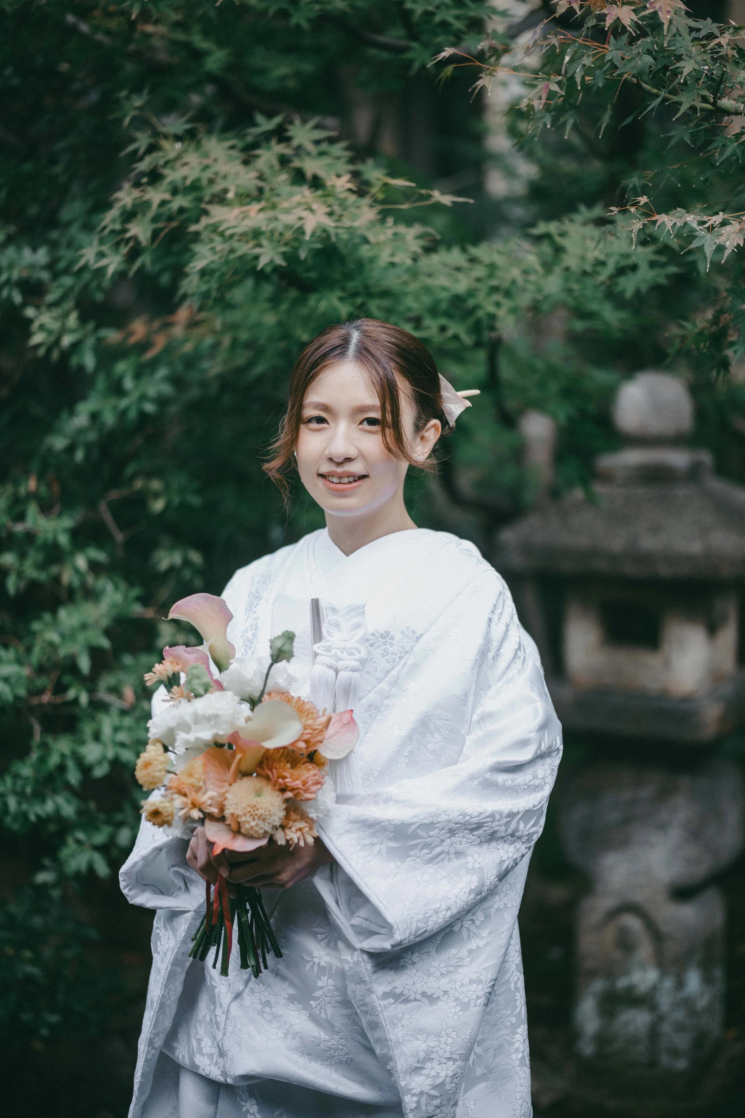 A young woman in a white traditional Japanese wedding kimono holding a bouquet of flowers, standing outdoors surrounded by green foliage and a stone lantern in the background.