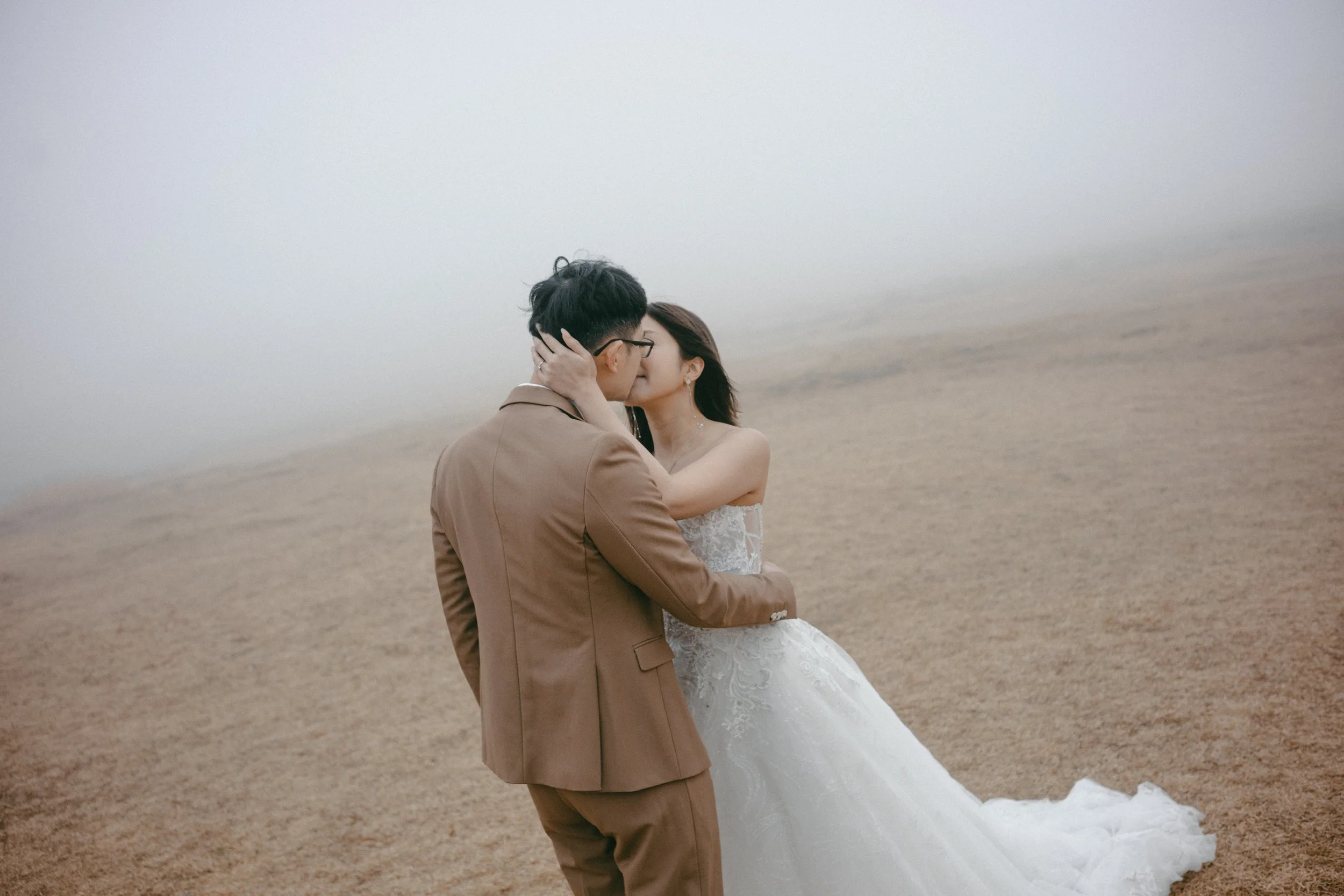 A couple dressed in wedding attire sharing a kiss on a foggy, deserted beach.