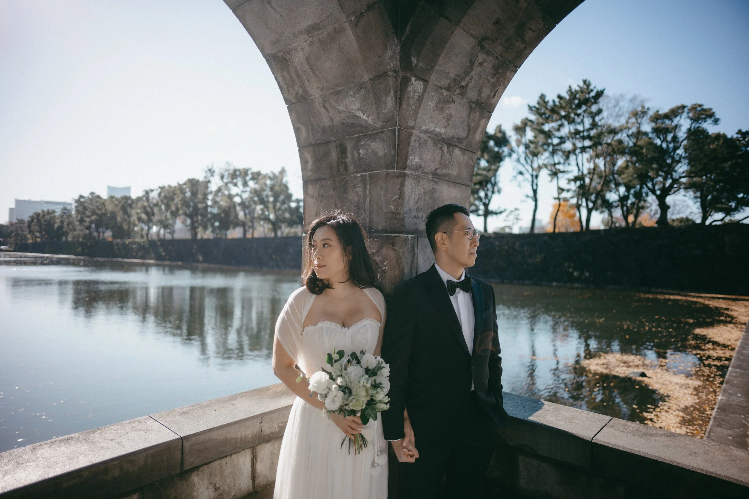 A bride and groom holding hands against an arch bridge near a river, with trees and buildings in the background.