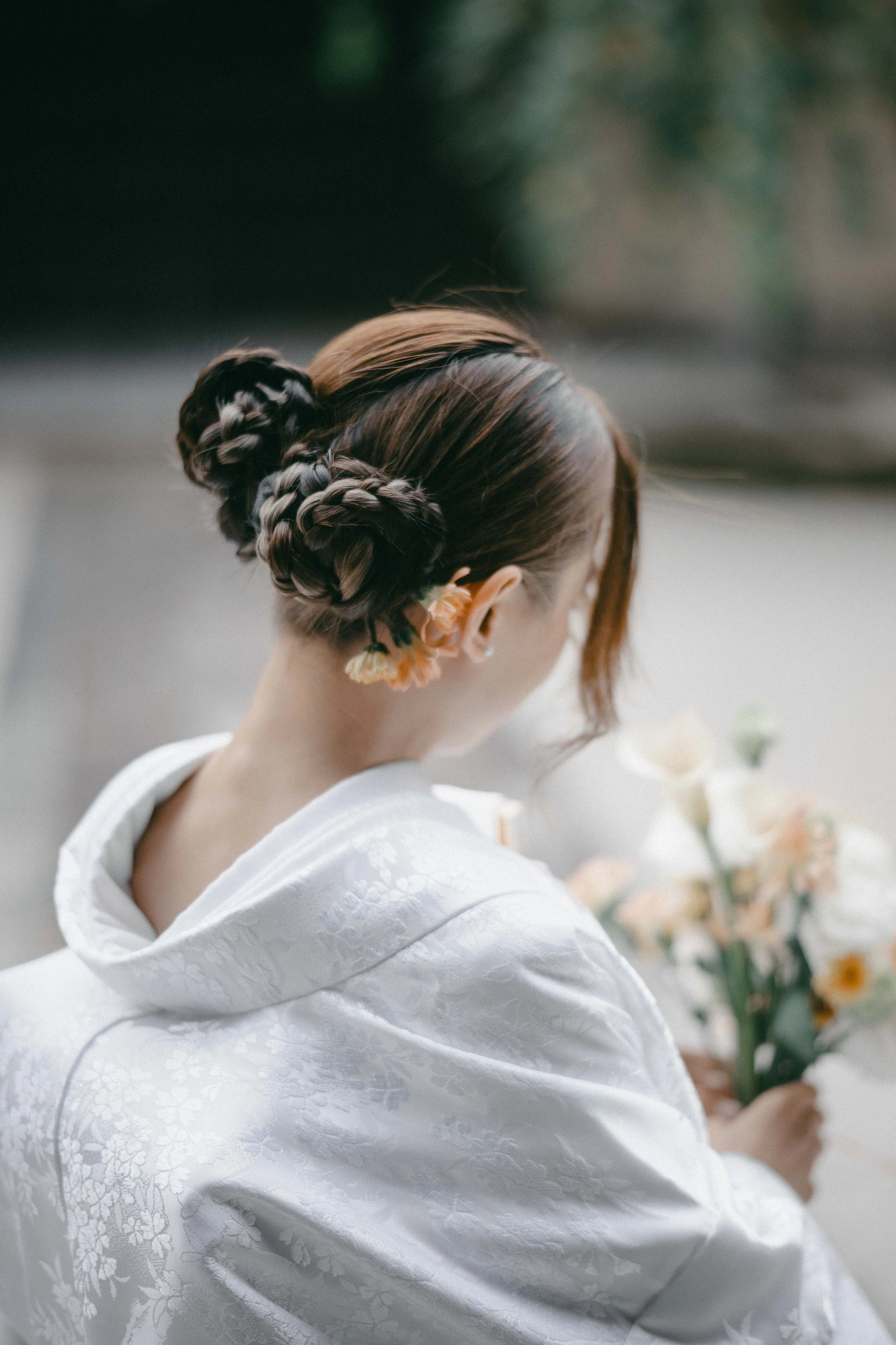 Woman with a braided updo hairstyle holding a bouquet of flowers, wearing a white garment with floral embroidery, seen from the back.