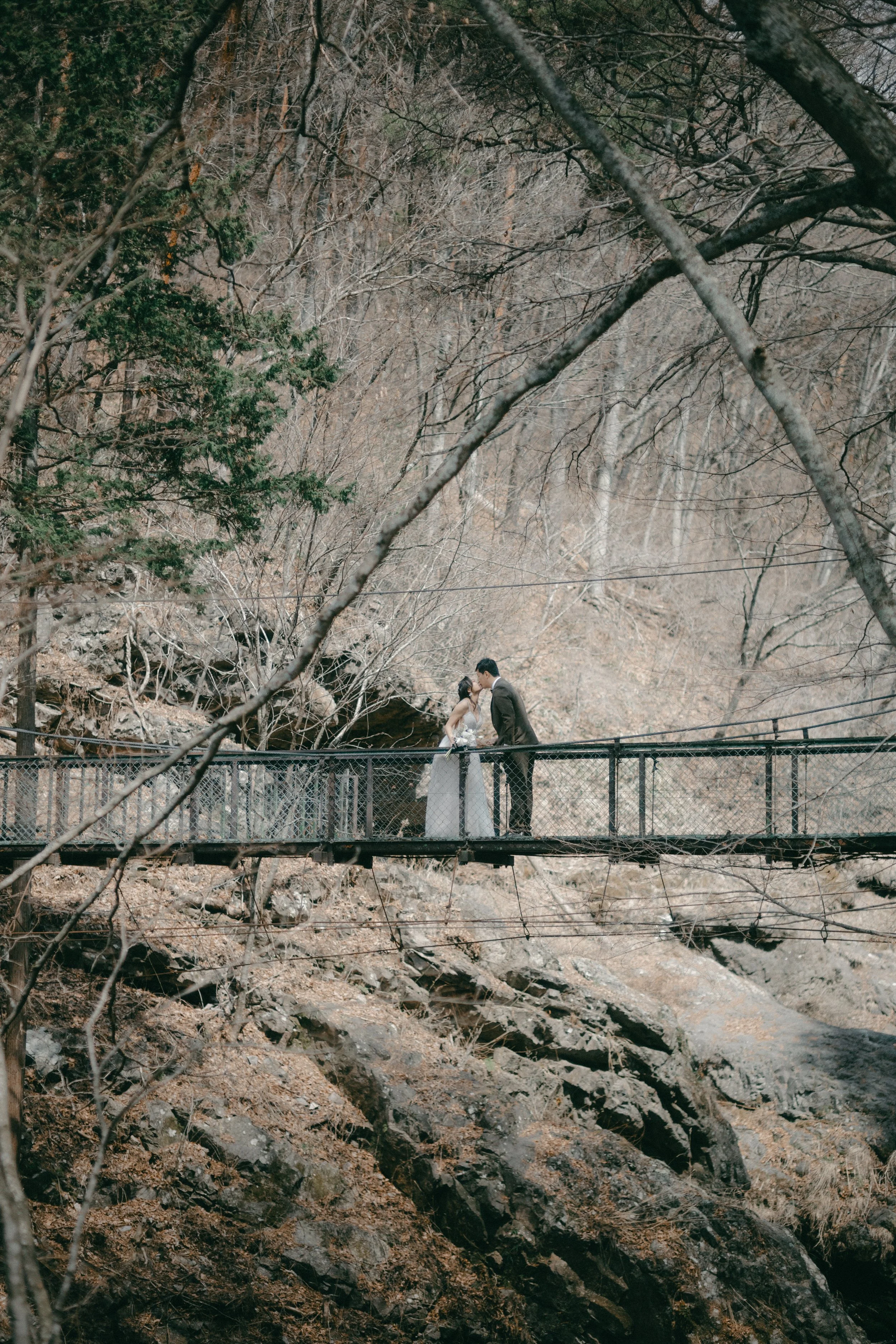 A couple dressed in wedding attire stands on a narrow footbridge in a forest, sharing a romantic moment as they lean in for a kiss.