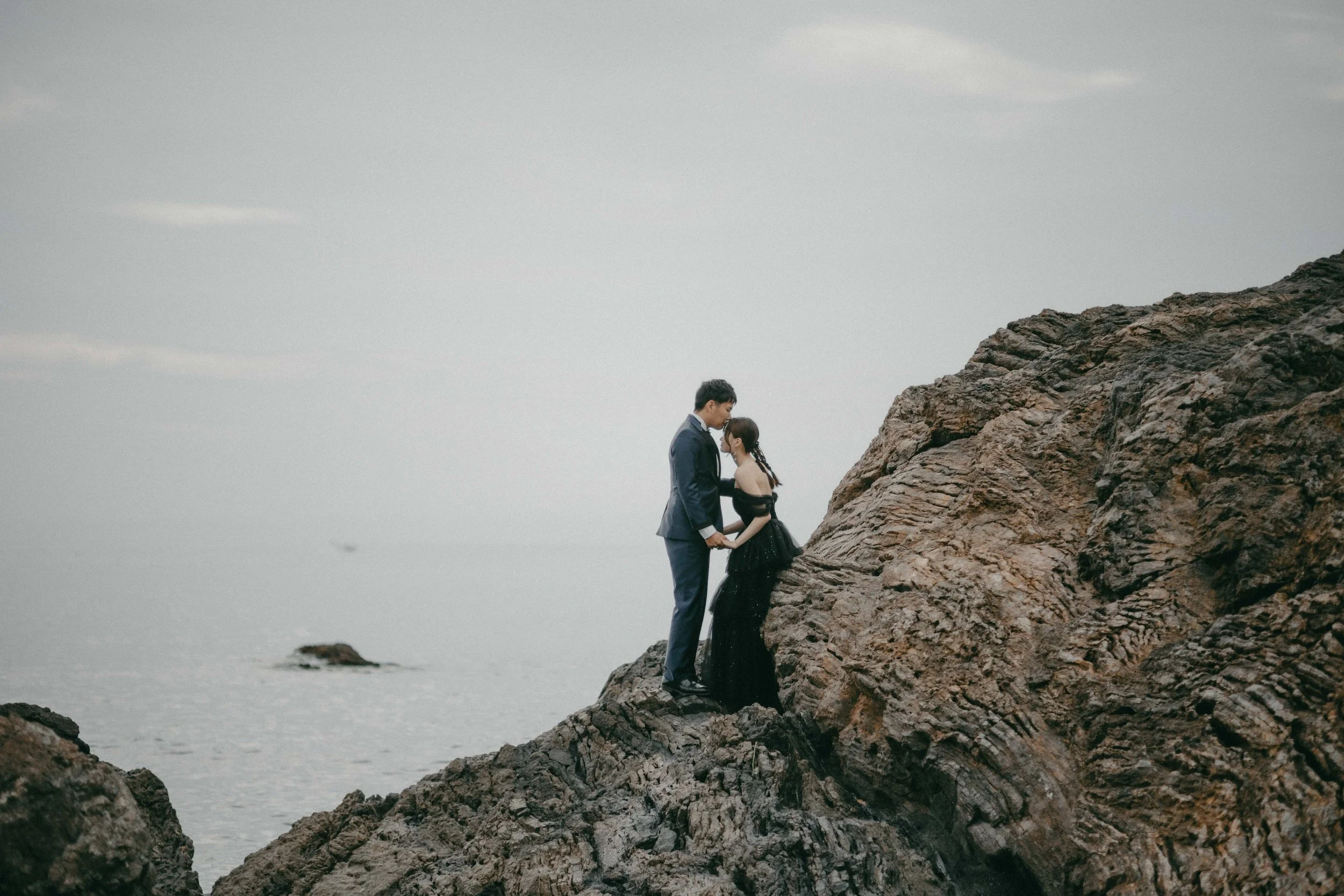 A man and woman in formal attire holding hands on large rocks by the ocean, with a cloudy sky above.