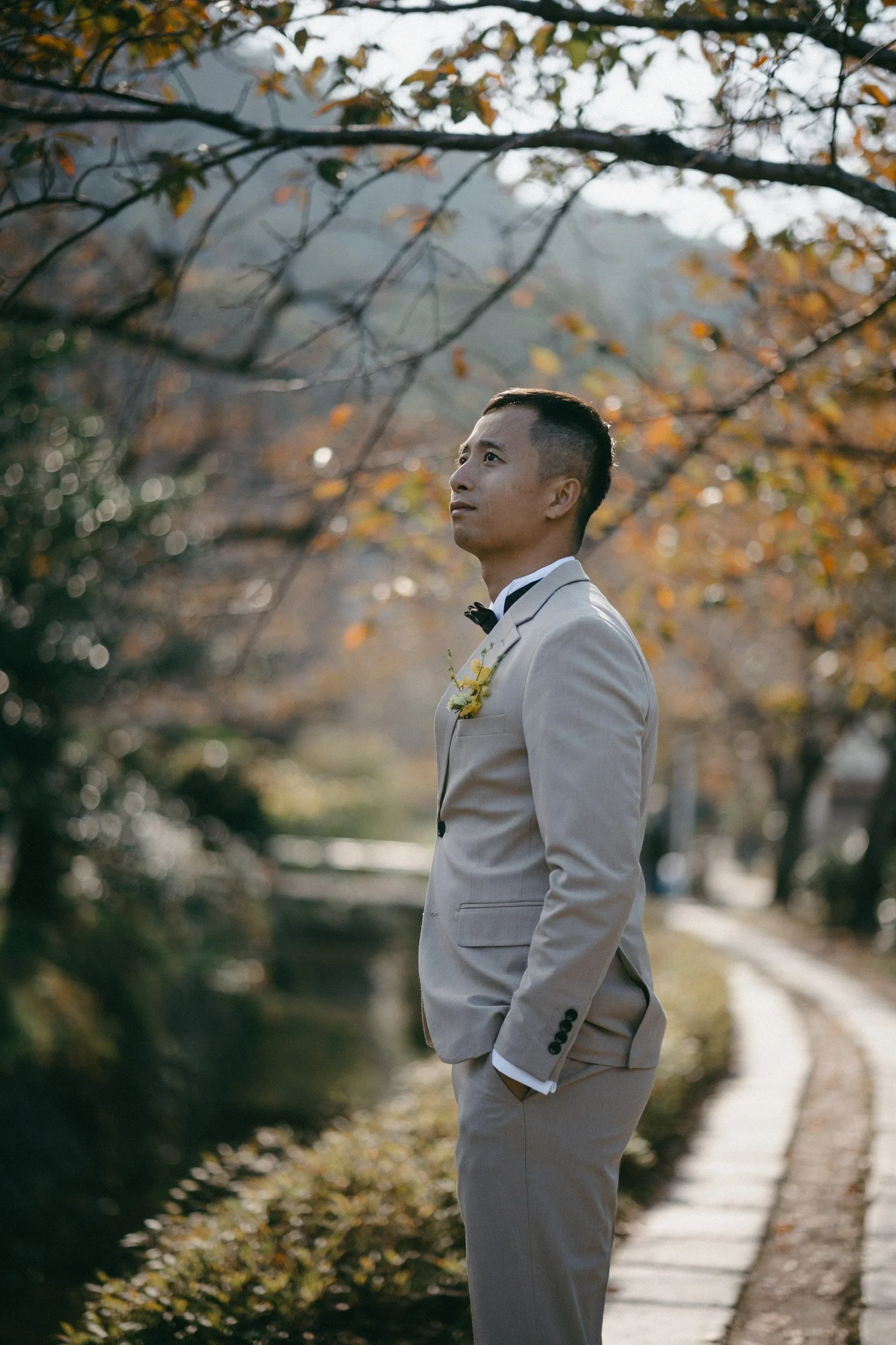 A groom in a light grey suit standing outdoors on a stone path among autumn trees, looking into the distance.