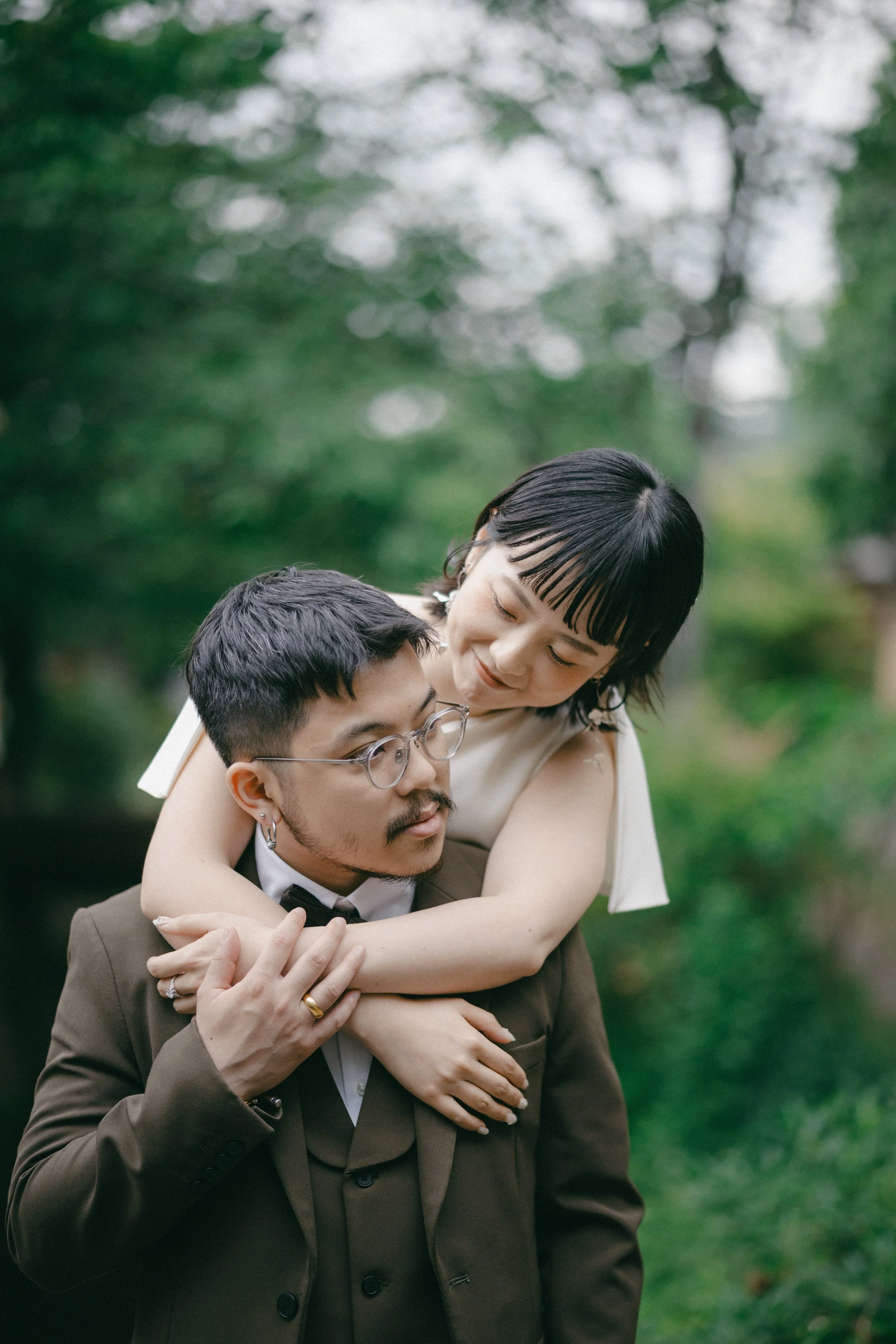 A couple outdoors with the woman on the man's shoulders, smiling at each other, surrounded by green trees.