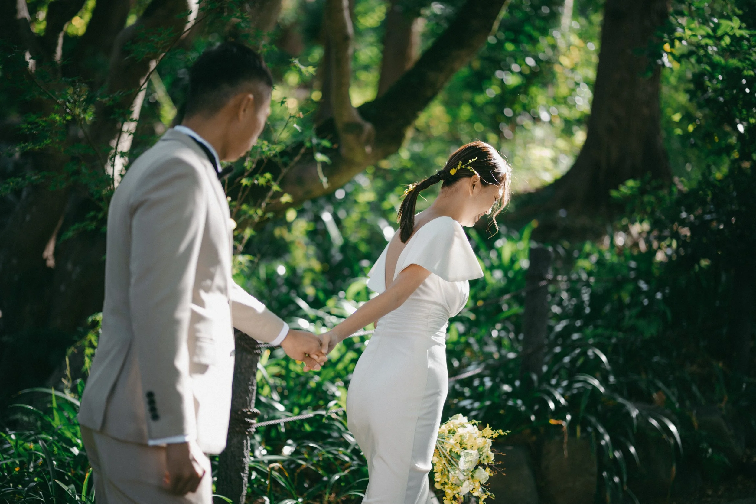 A couple in wedding attire holding hands in a lush green outdoor setting with sunlight filtering through trees.