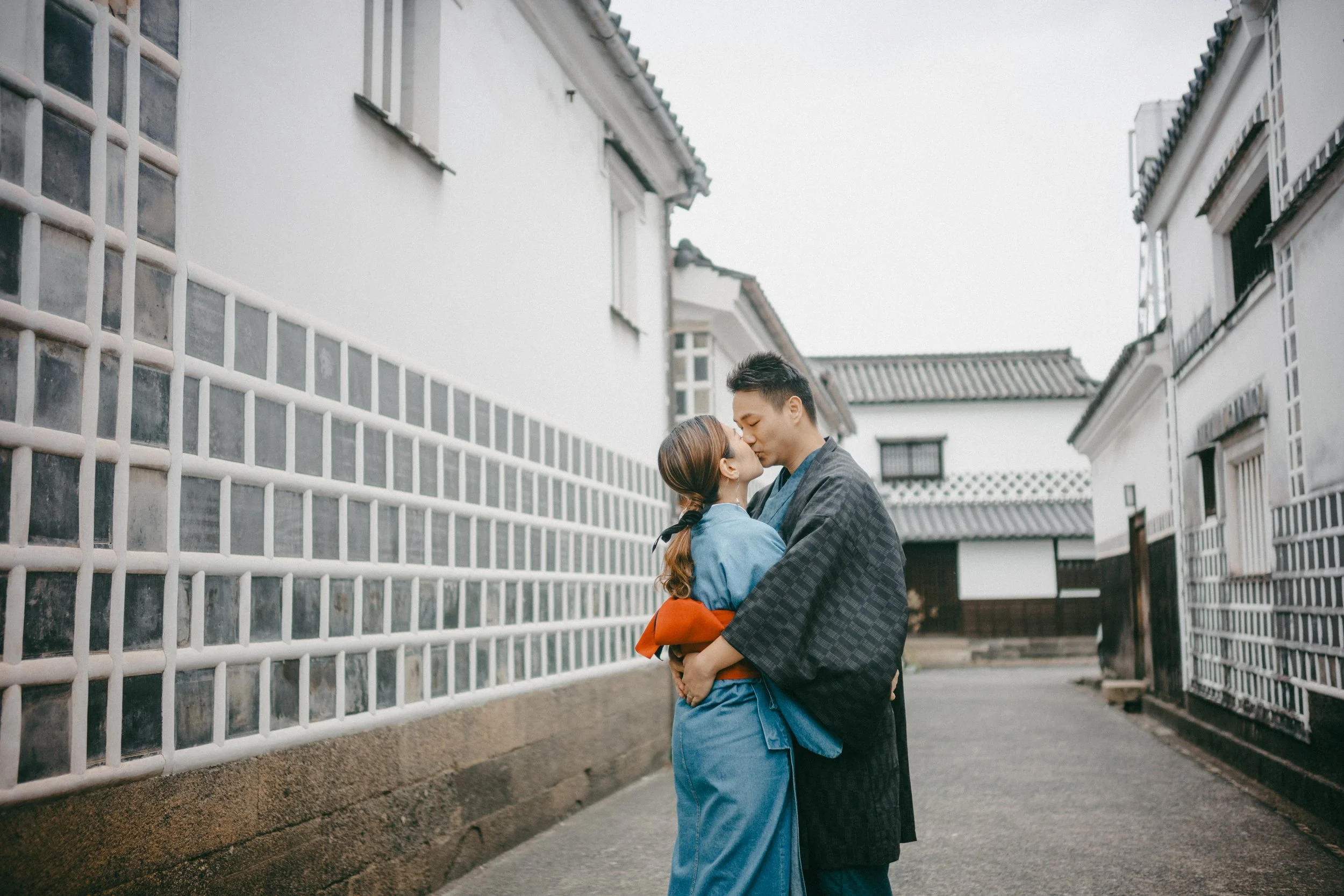 A couple dressed in traditional Japanese clothing sharing a kiss on a quiet street with white walls and tiled roofs.