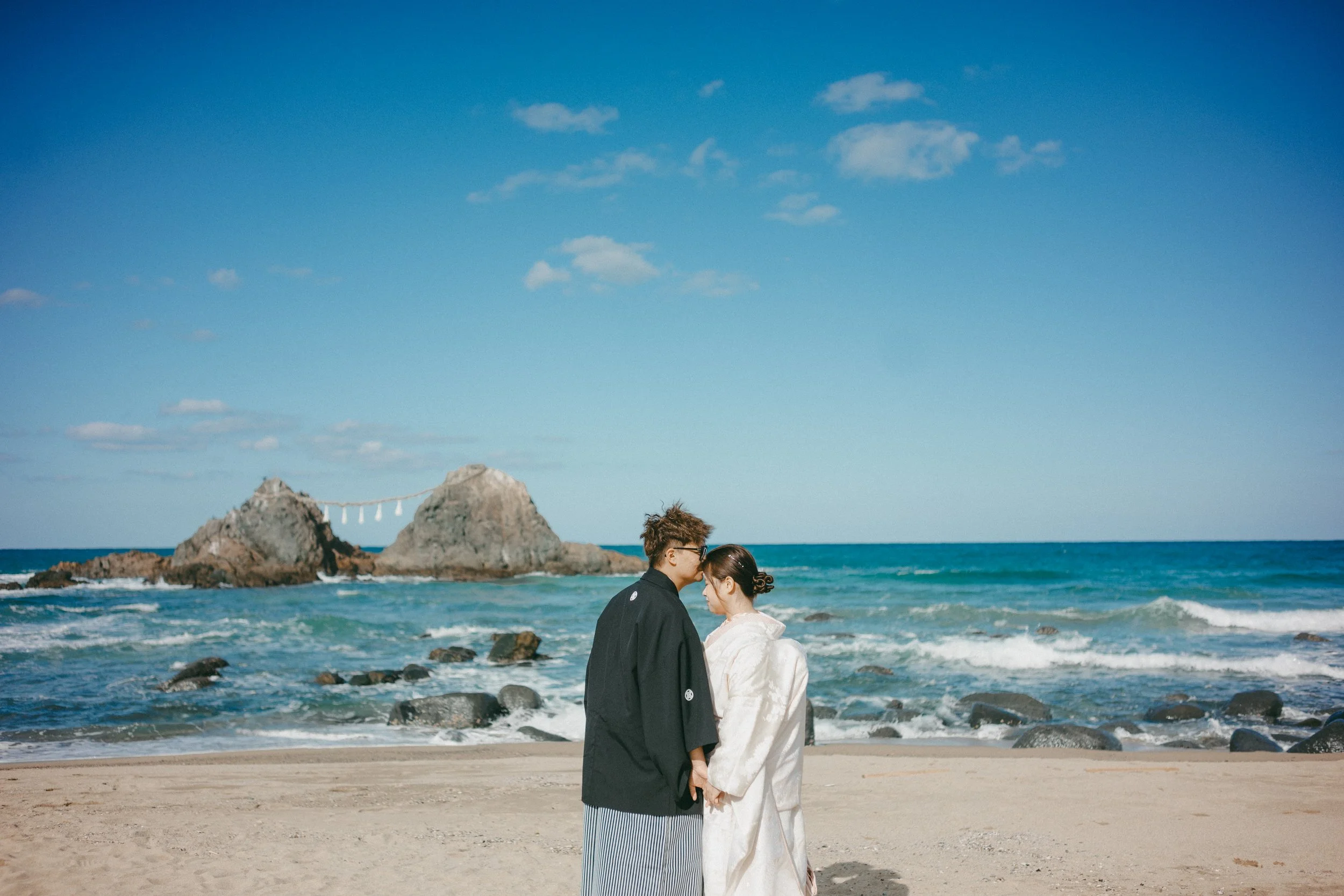 A couple dressed in traditional Japanese wedding attire holding hands and touching foreheads on a beach with rocks and a large rock formation in the background, under a blue sky with some clouds.