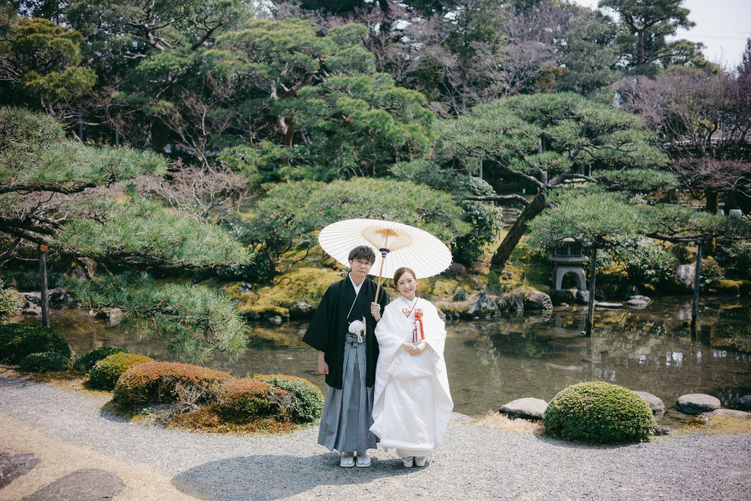 A young couple dressed in traditional Japanese wedding attire stands on a path in a Japanese garden, holding a white parasol. The man wears a black kimono with striped hakama, and the woman wears a white kimono with red accents, smiling at the camera