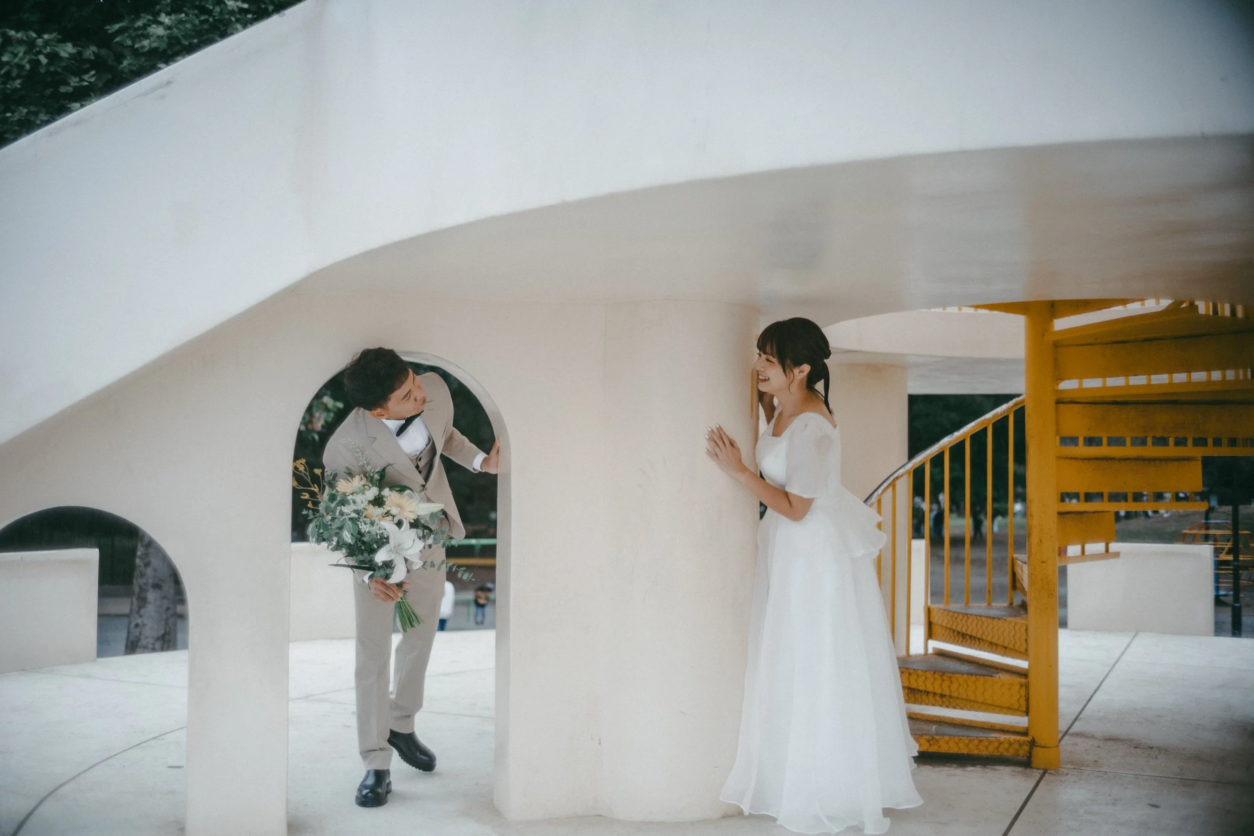 A bride and groom in wedding attire smiling at each other by a white curved wall with a yellow spiral staircase in the background