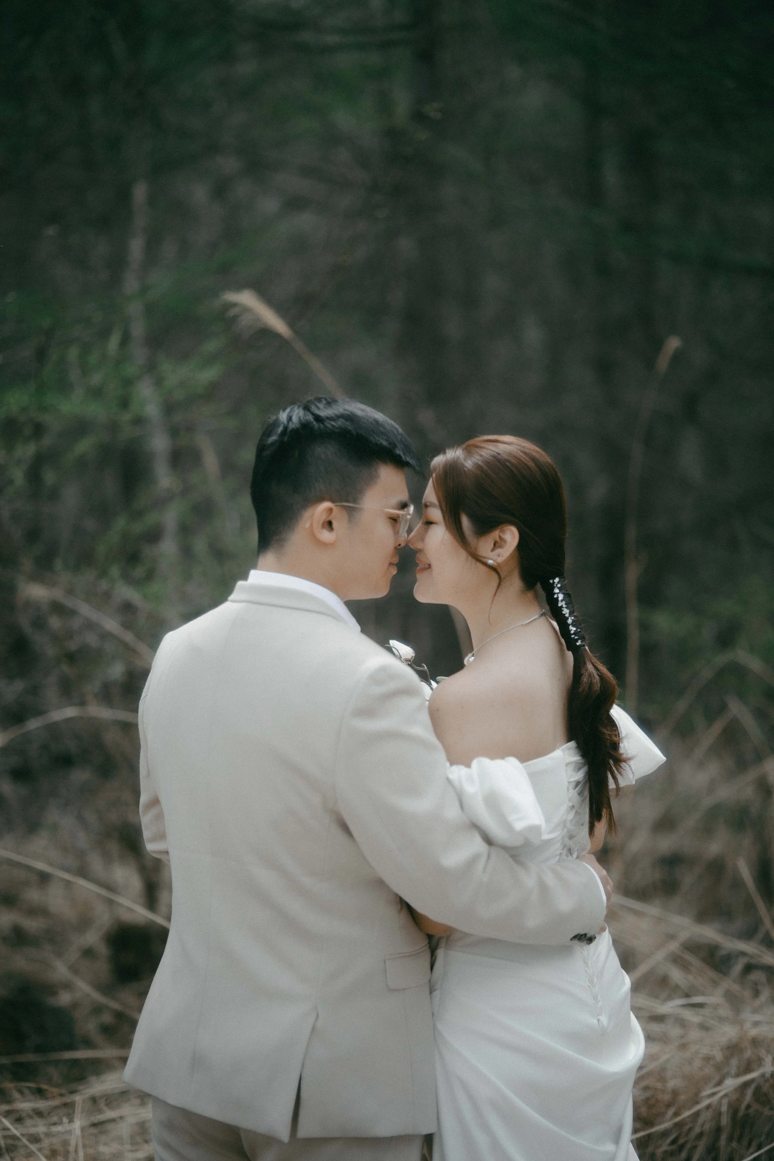 A couple in white wedding attire standing close together outdoors, foreheads touching, with beaming smiles in a natural setting with blurred greenery in the background.