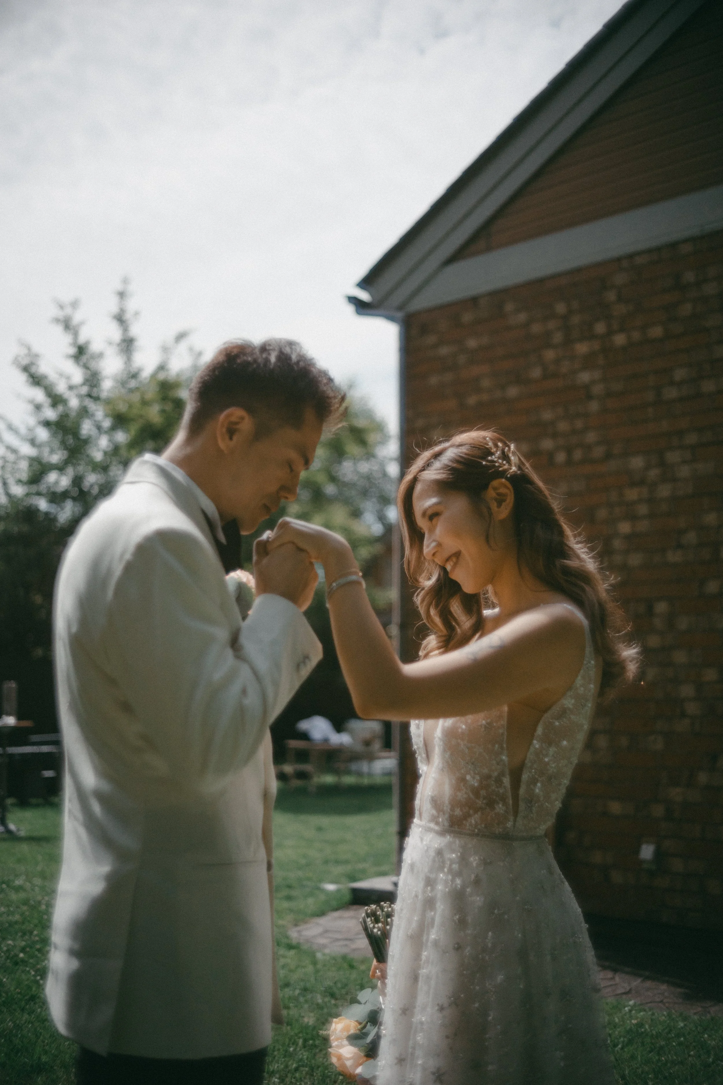 A bride and groom share an intimate moment outside on their wedding day, smiling and holding hands, with a brick house and greenery in the background.