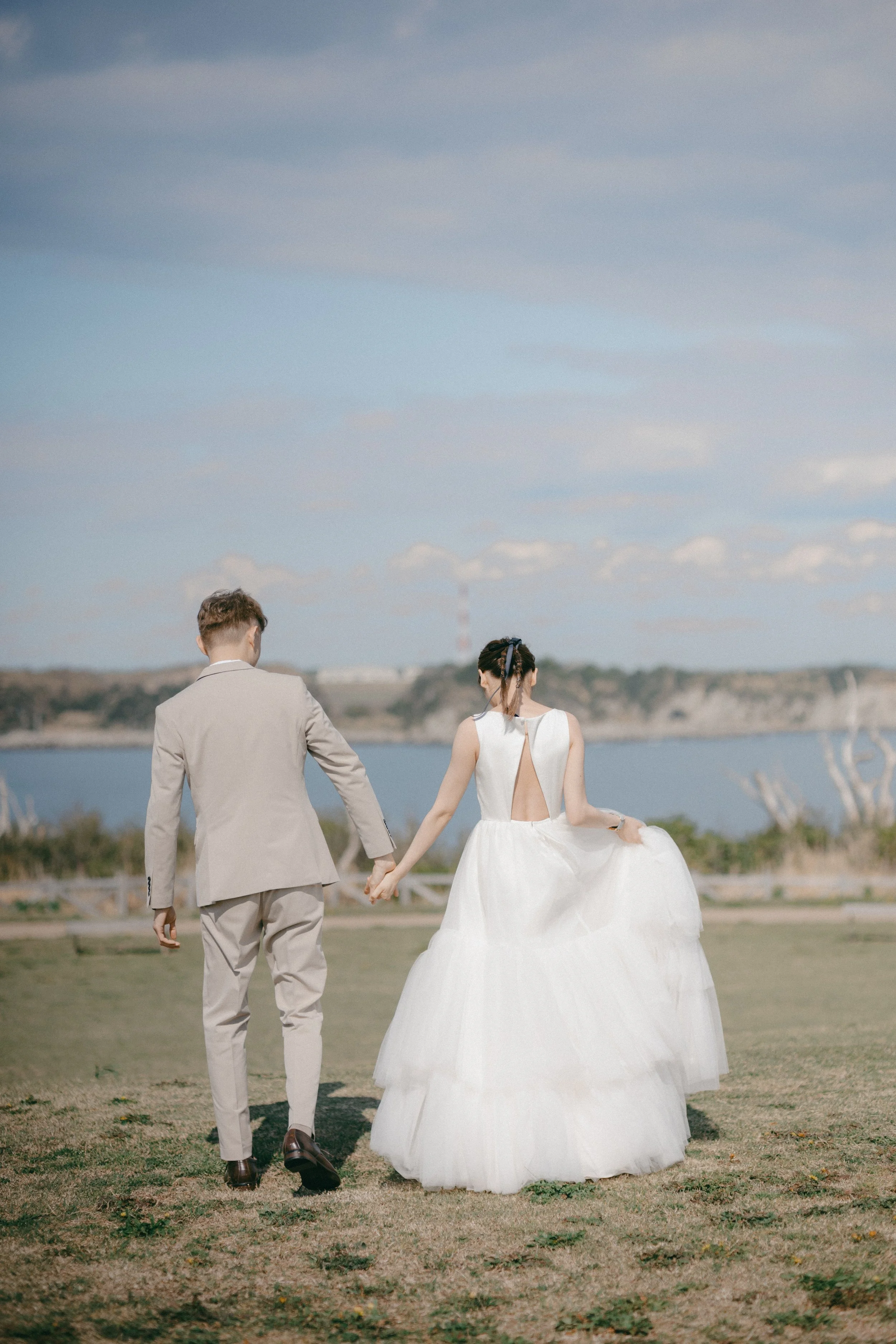 Couple in wedding attire walking hand in hand on a grassy field near a body of water, with a distant shoreline and a cloudy sky in the background.