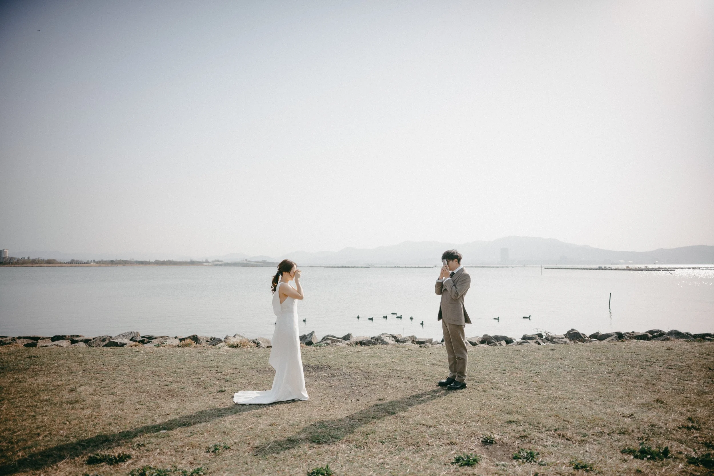 A bride in a white wedding dress and a groom in a beige suit standing apart facing each other on a grassy area near a body of water, with ducks swimming in the water and mountains in the distance under a clear sky.