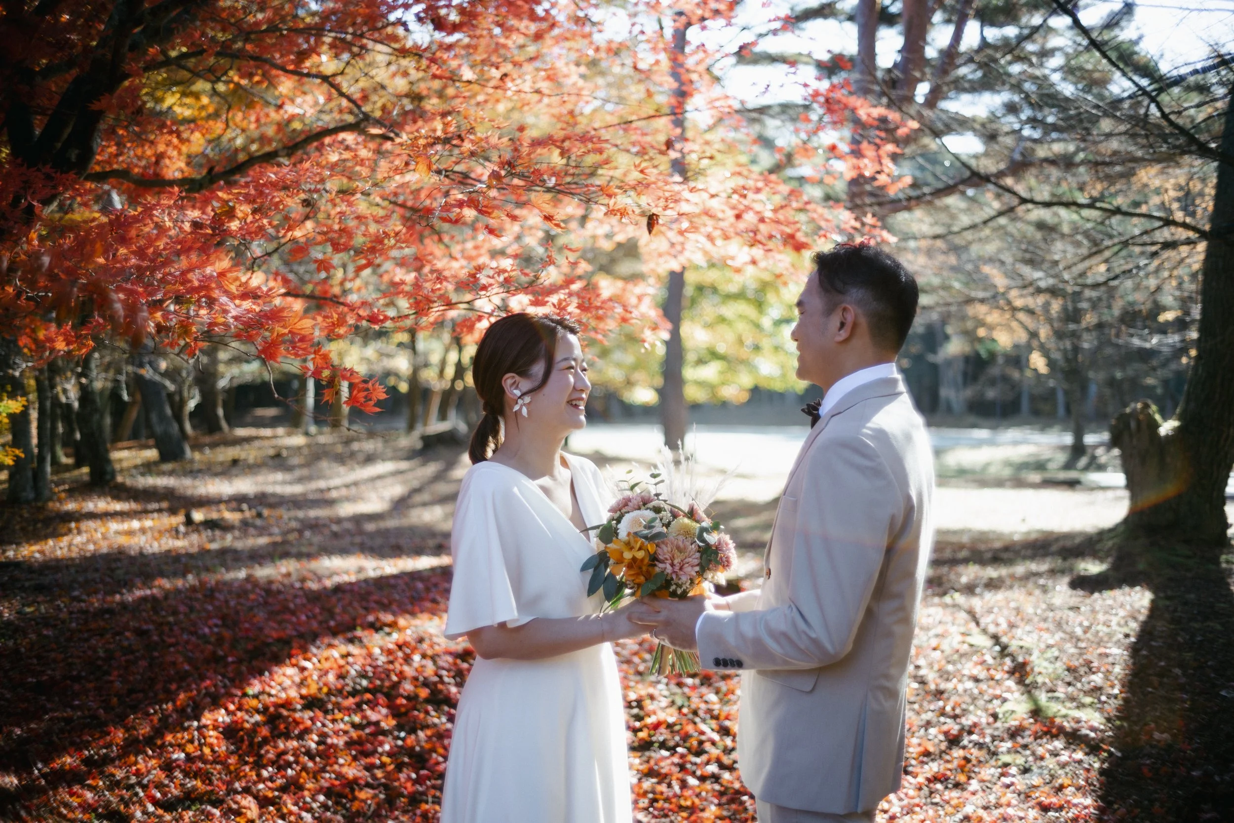 A bride and groom stand face to face outdoors during autumn, smiling at each other, with colorful fall leaves on trees and ground; the bride holds a bouquet of flowers.