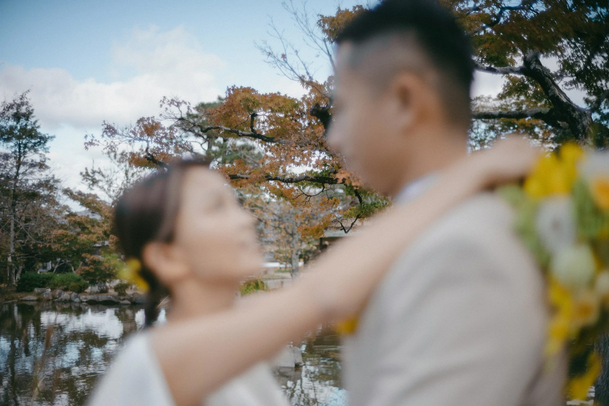 A couple in outdoor autumn setting, with trees and a pond in the background, embracing each other, faces touching, in a romantic moment.