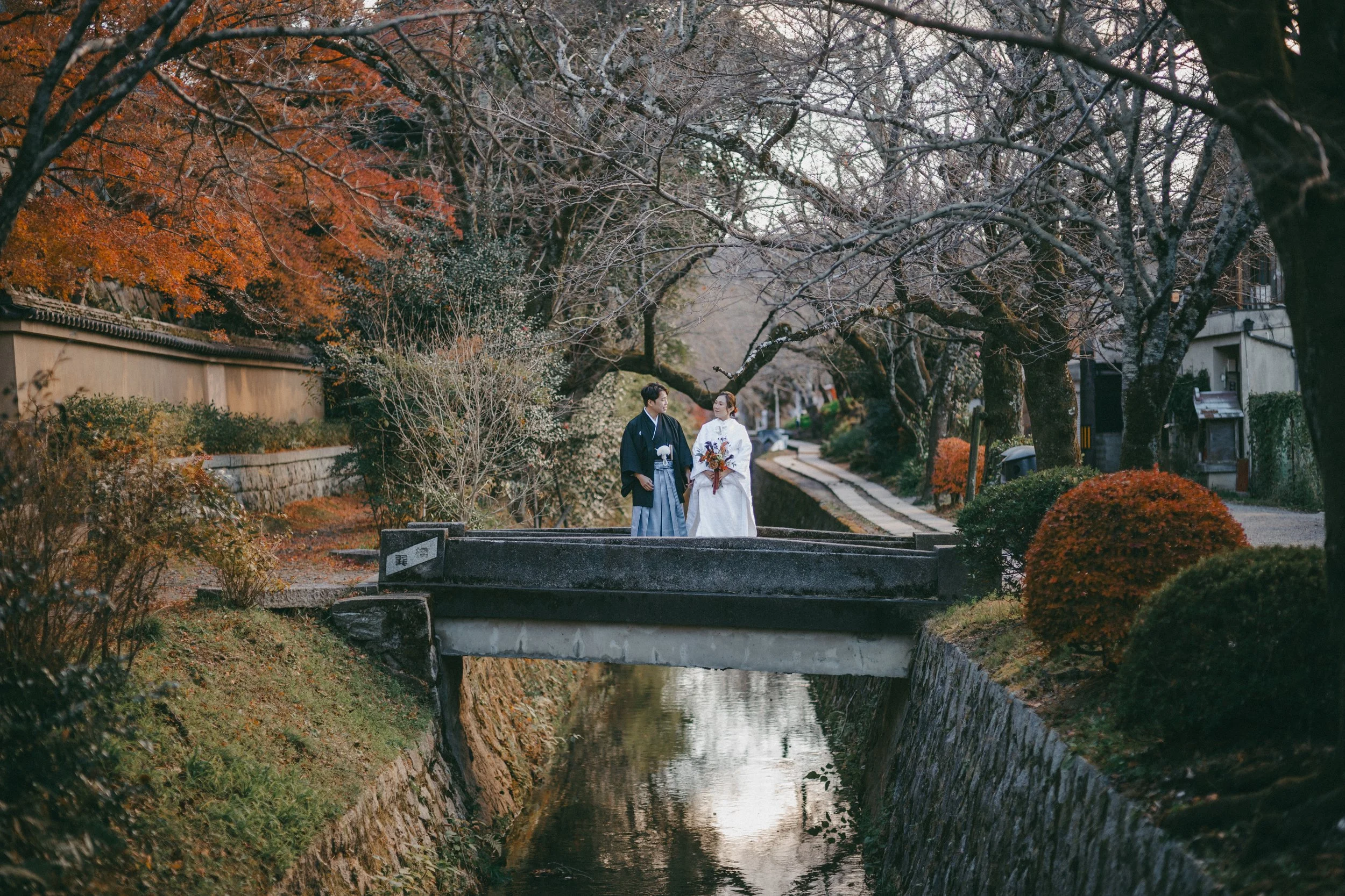 A couple dressed in traditional Japanese wedding attire standing on a small bridge over a narrow canal, surrounded by autumn trees with orange and red leaves.