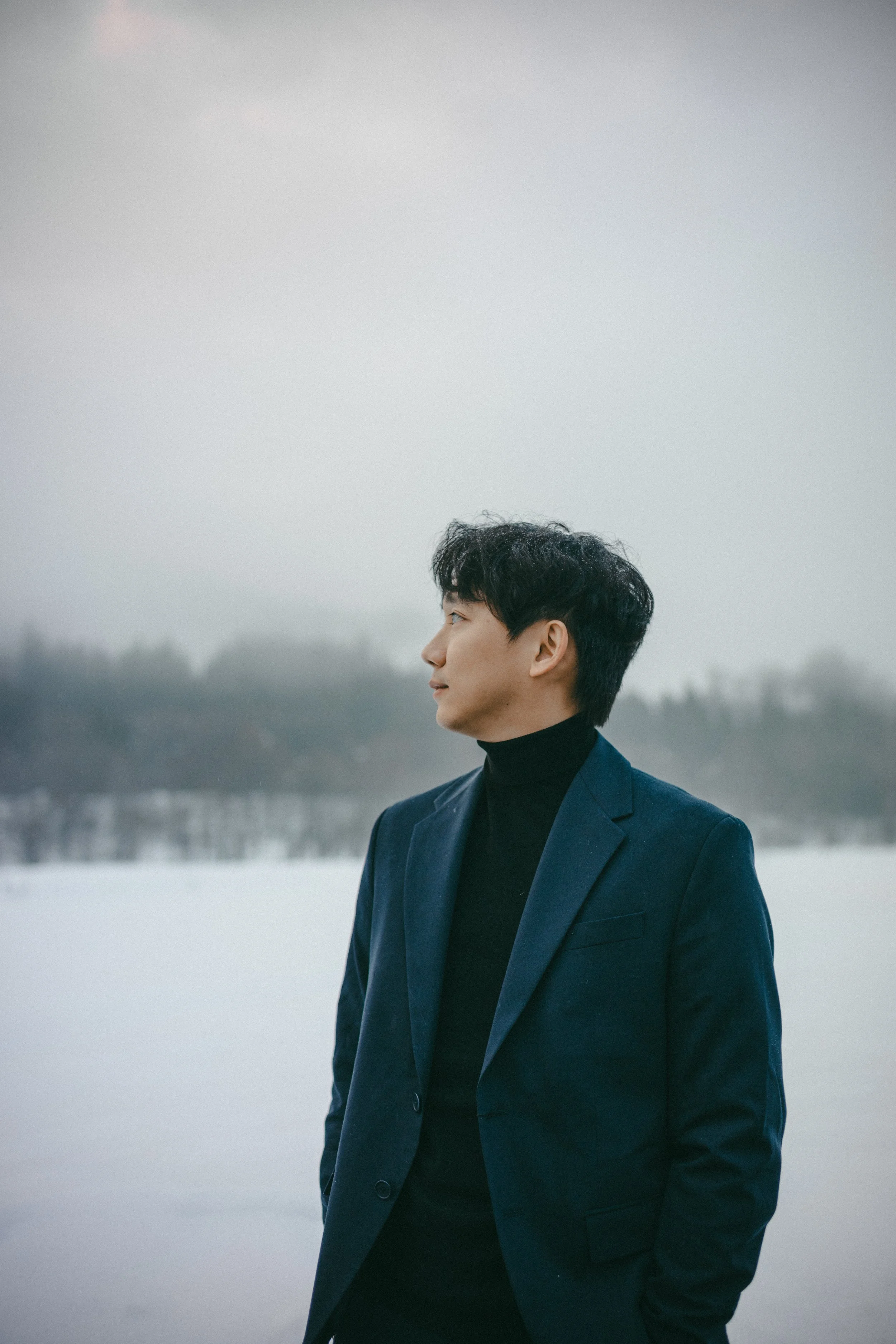 A man in a navy blazer and black turtleneck stands outdoors near a frozen lake or river, with a cloudy sky and distant trees in the background.