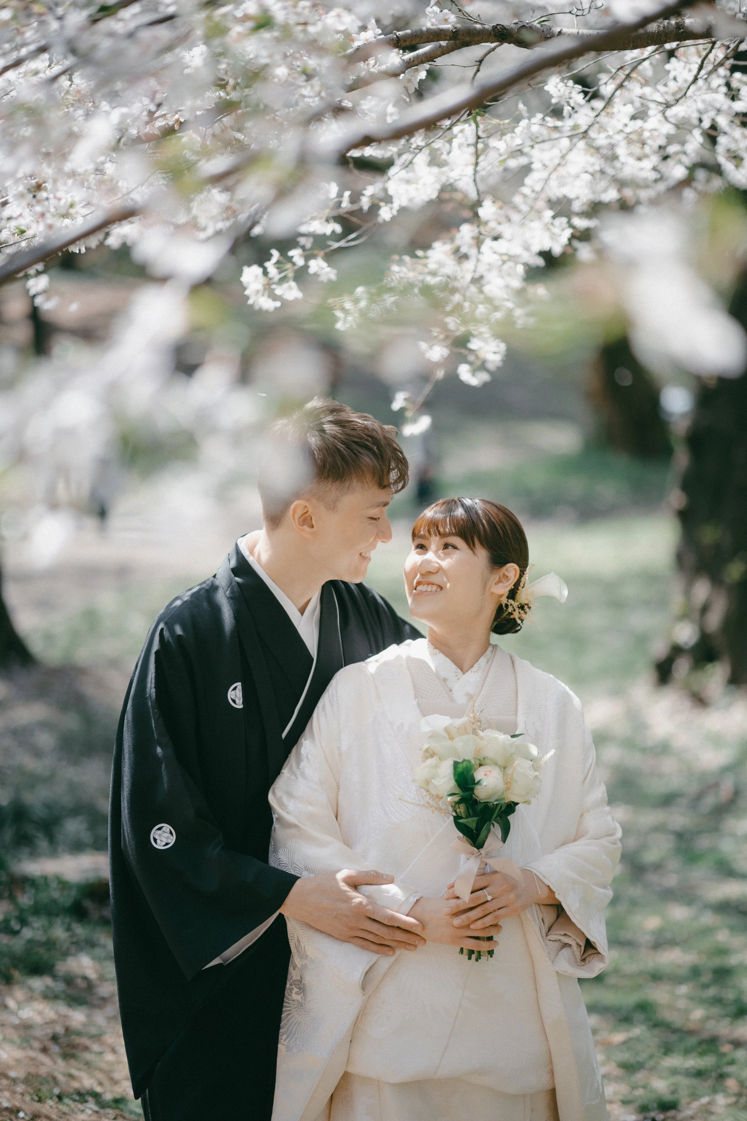 A smiling couple dressed in traditional Japanese wedding attire stands close under blooming cherry blossom trees during daytime. The bride holds a bouquet of white roses.
