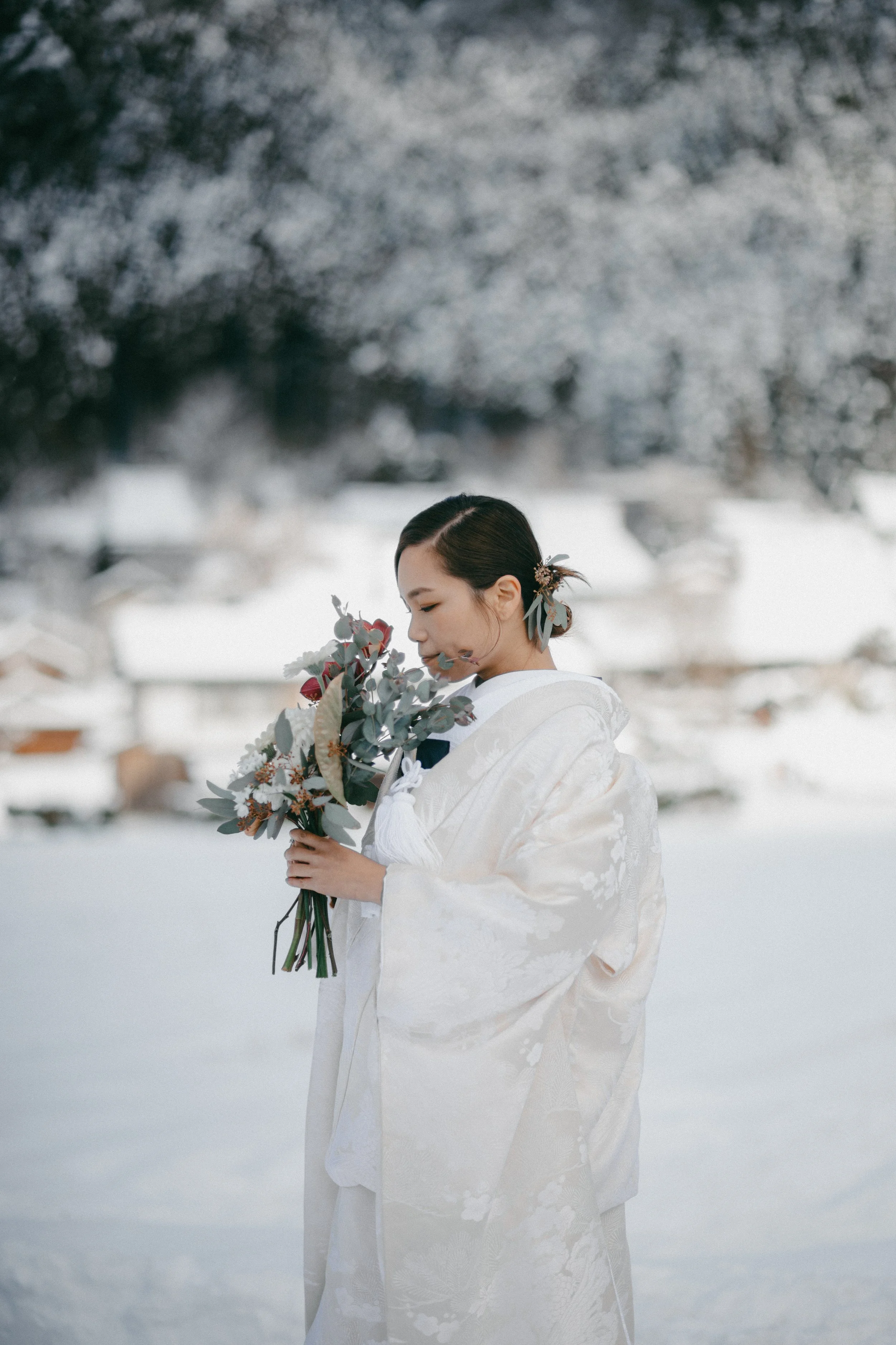 A woman wearing a traditional white wedding kimono holding a bouquet of flowers outdoors in a snow-covered landscape.