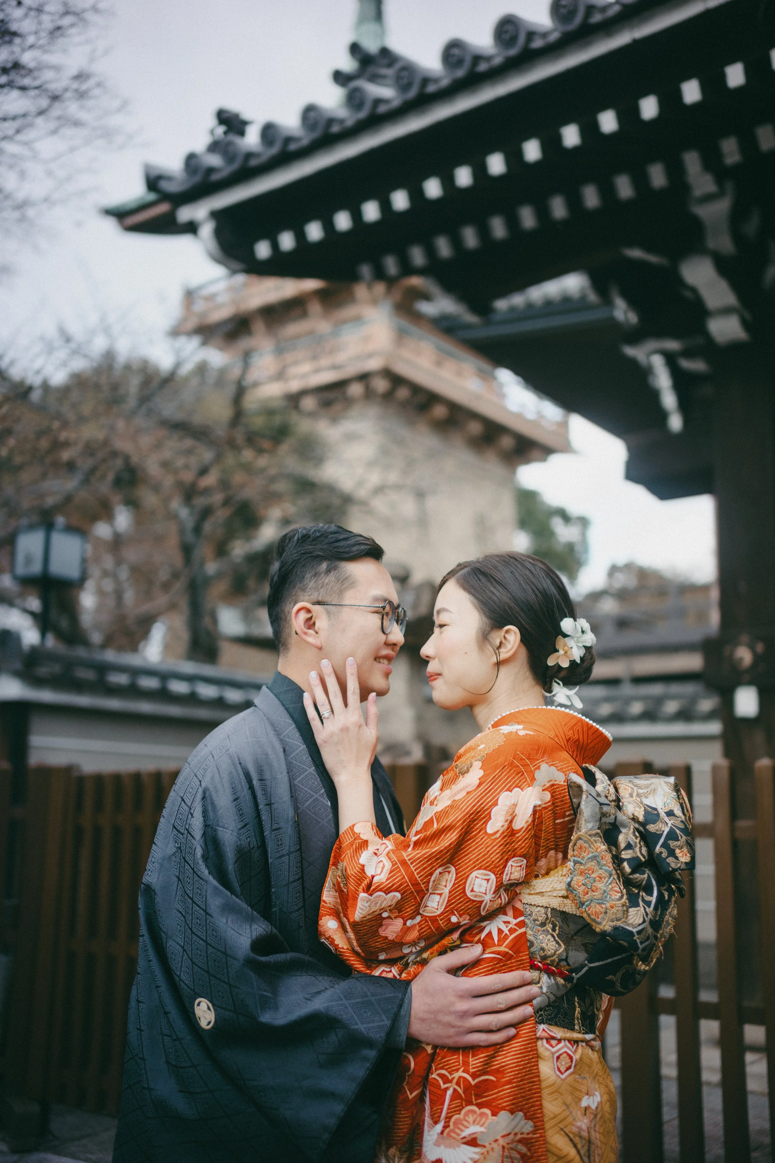 A couple dressed in traditional Japanese kimono and kimono jacket embracing and touching foreheads in front of a historical Japanese temple or shrine.