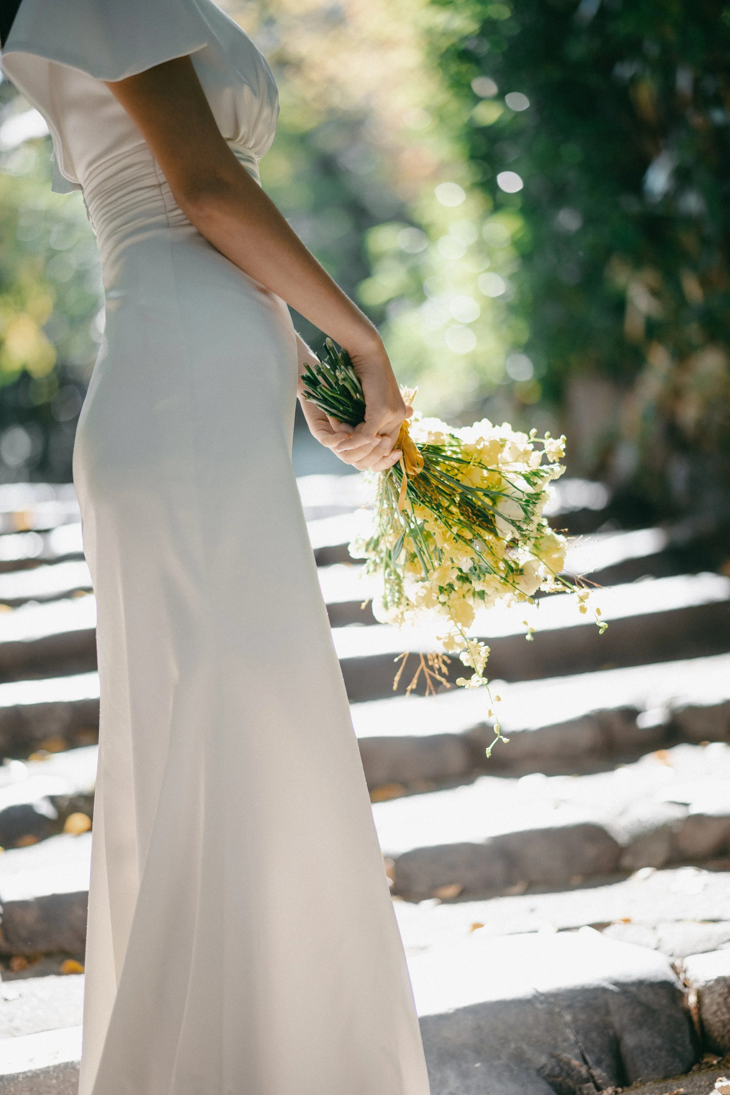 A woman in a white dress holding a bouquet of yellow flowers on a stone staircase outdoors.