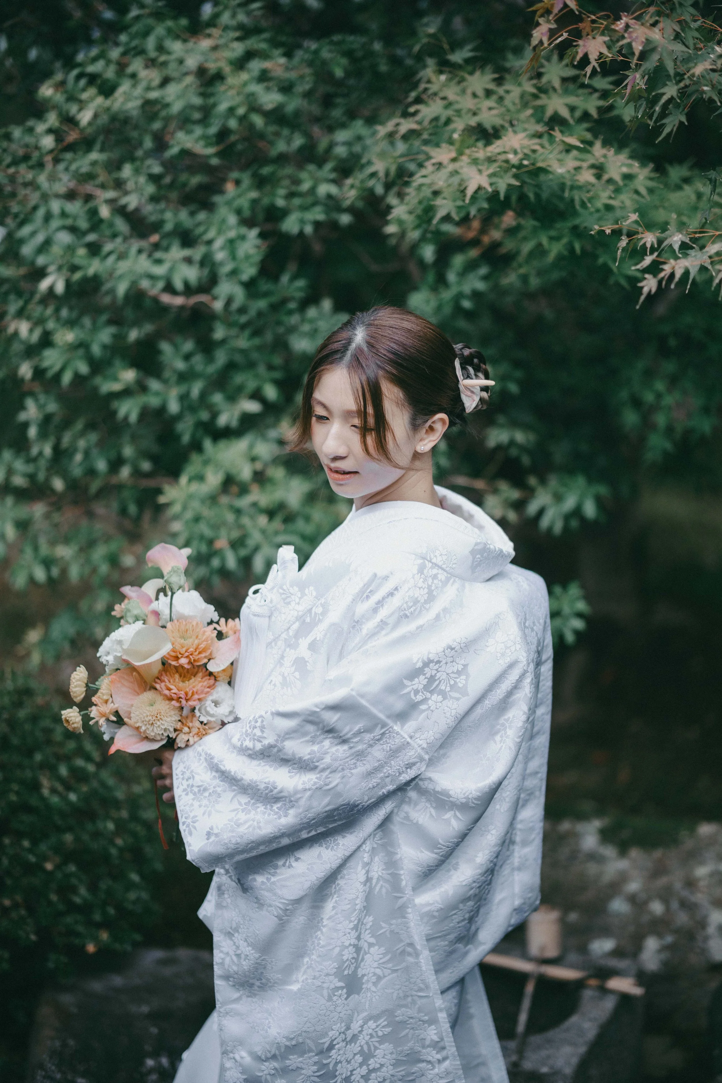 A woman dressed in a traditional white kimono holding a bouquet of flowers, standing in a lush garden with green foliage.
