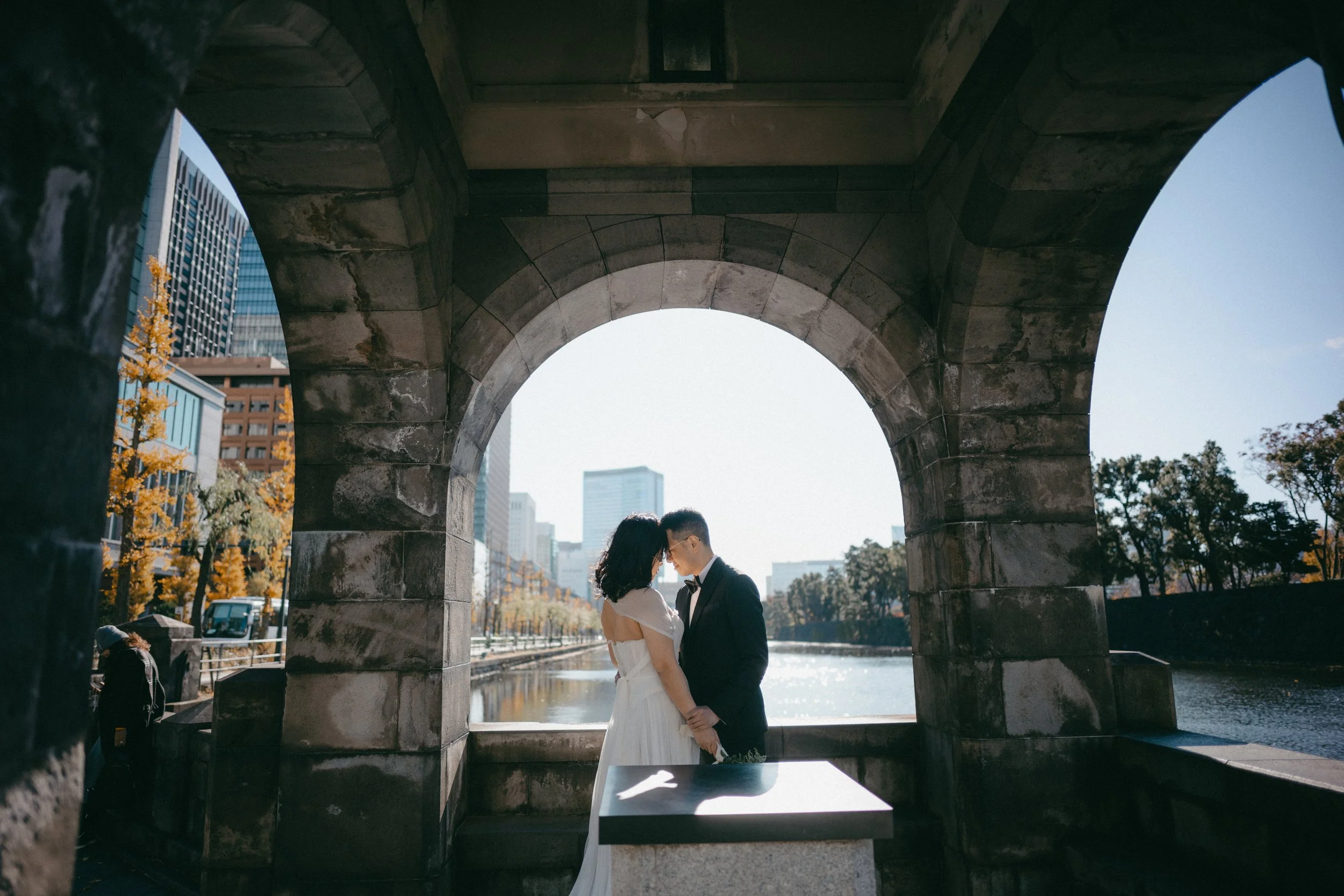 A couple in wedding attire, a woman in a white dress and a man in a black tuxedo, standing close together under a stone archway by a river in an urban area, with city buildings and autumn trees in the background.