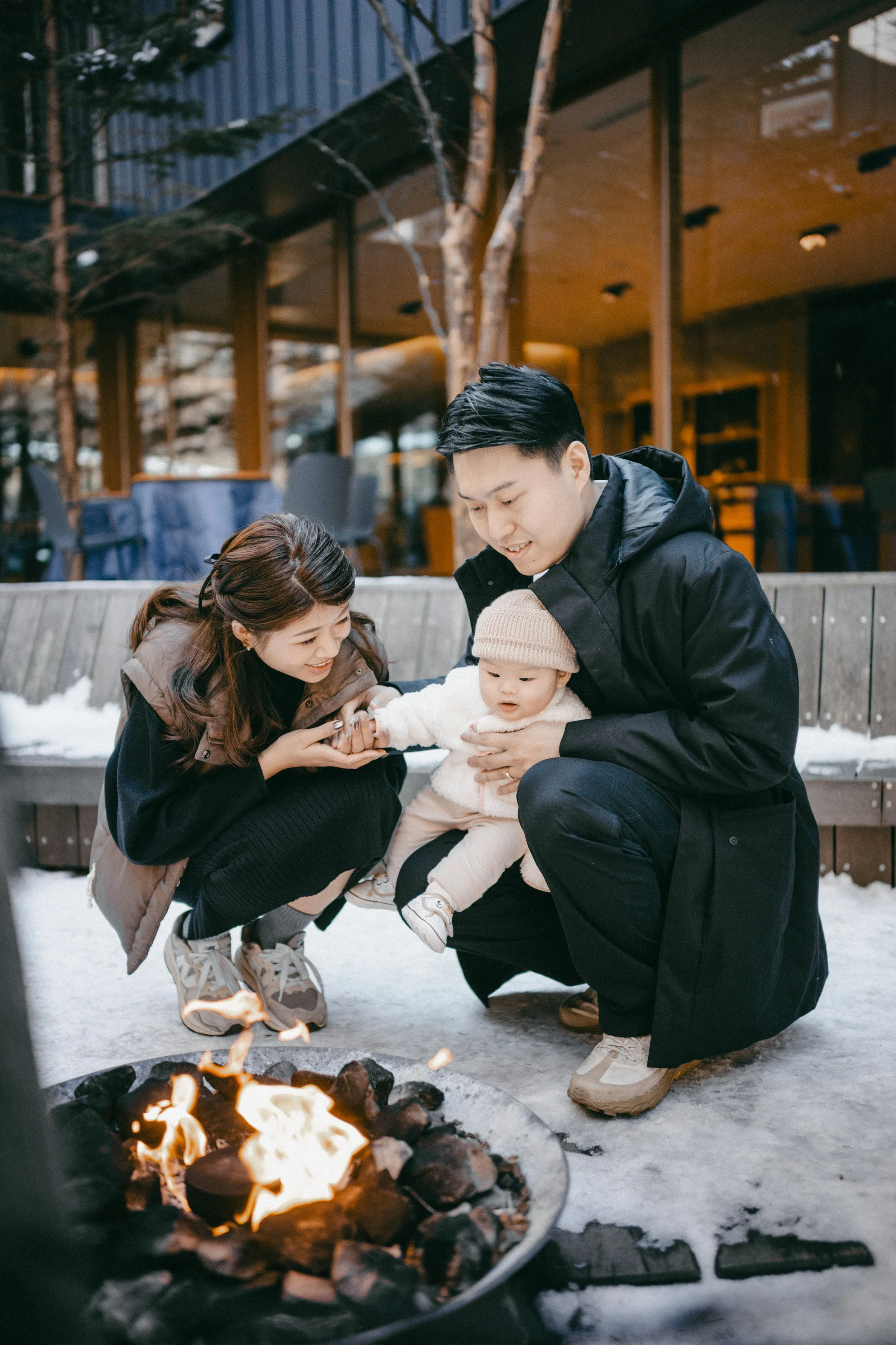 A family of three, including a woman, a man, and a baby, is gathered around a fire pit outside on snow-covered ground. The woman is crouching, holding the baby’s hand, and smiling, while the man is holding the baby, who is dressed in warm clothing an