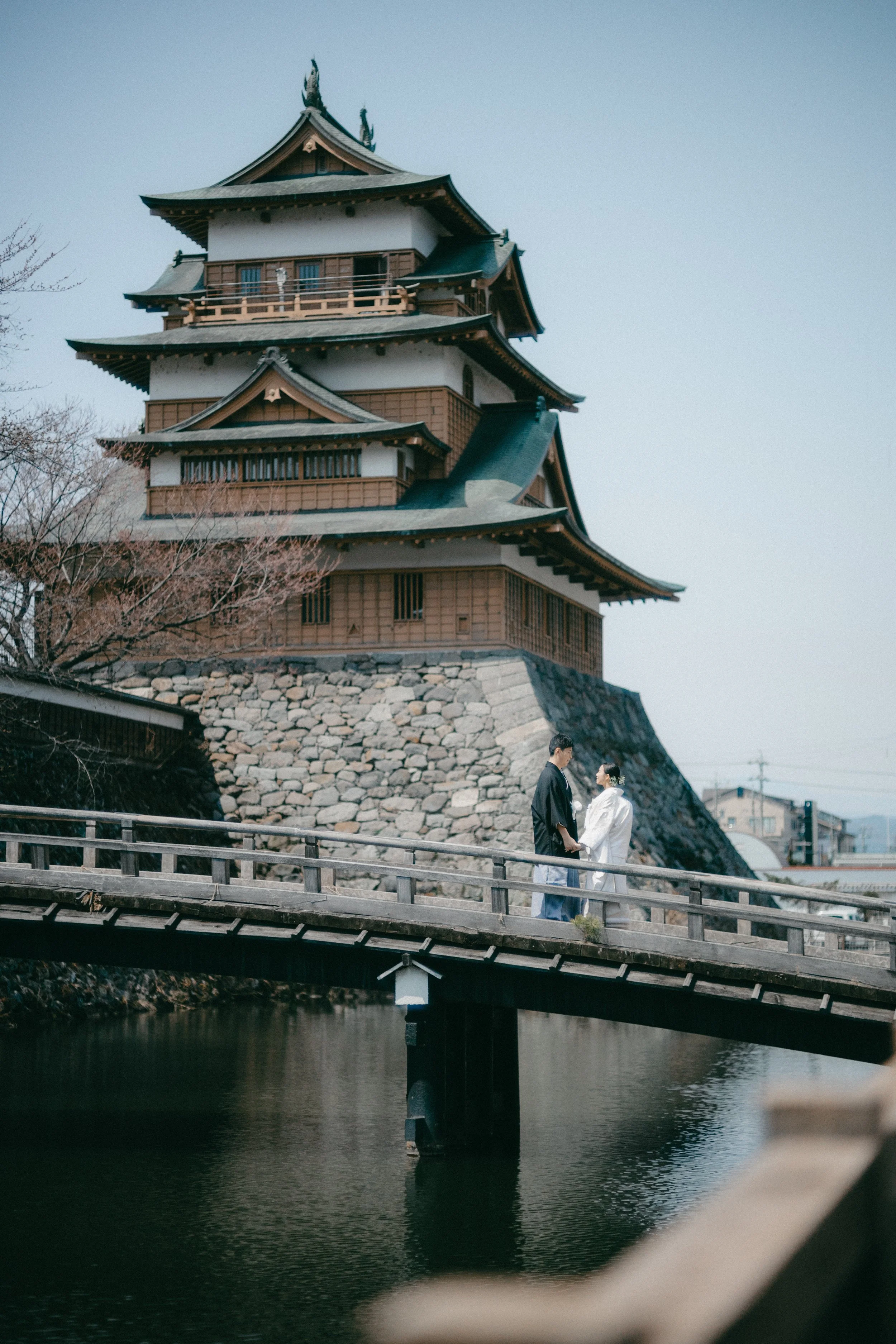 A couple dressed in traditional Japanese wedding attire holding hands and facing each other on a wooden bridge over a pond, with a traditional Japanese castle in the background.