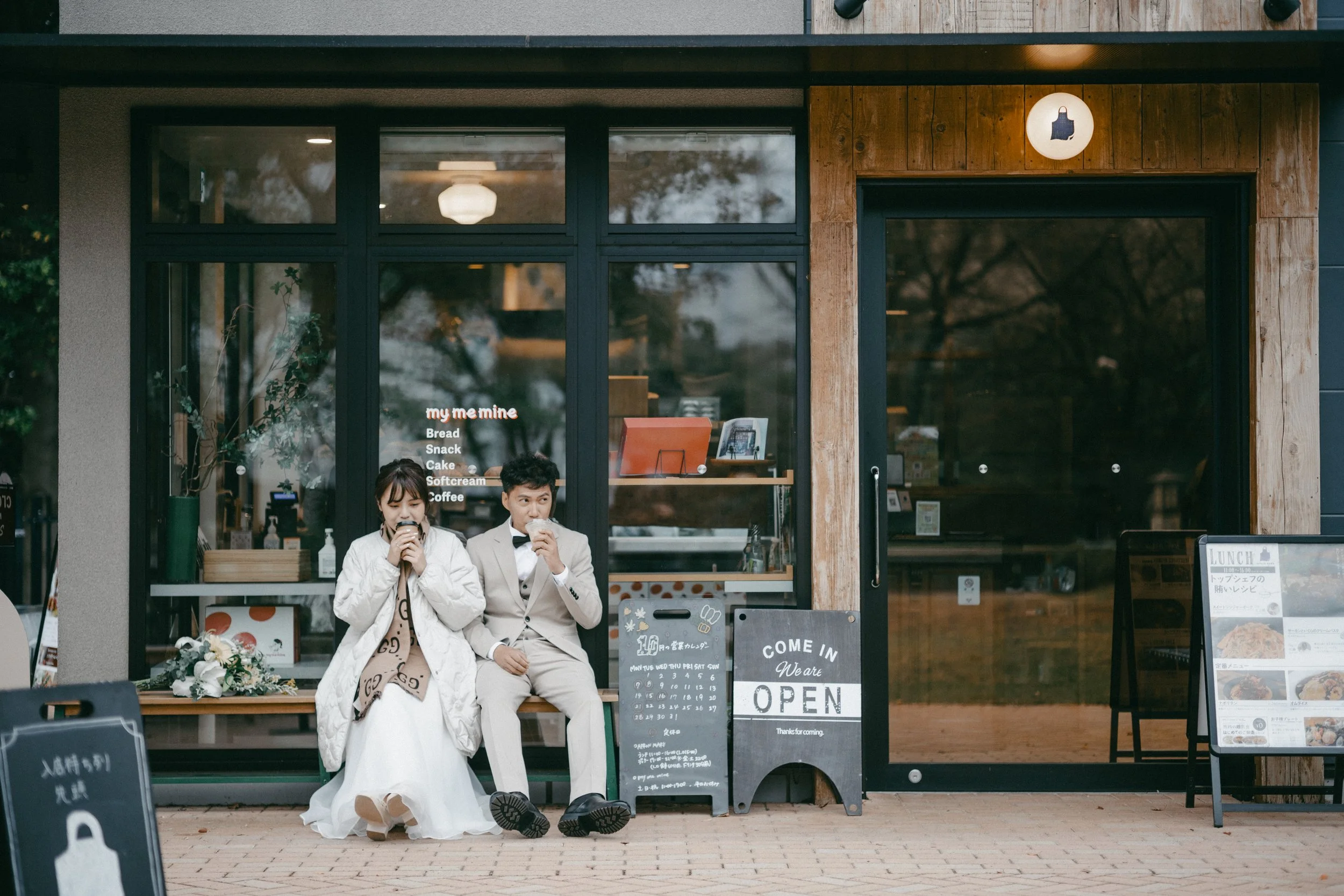 A young couple sitting outside a café, drinking coffee, with a storefront with large glass windows behind them.