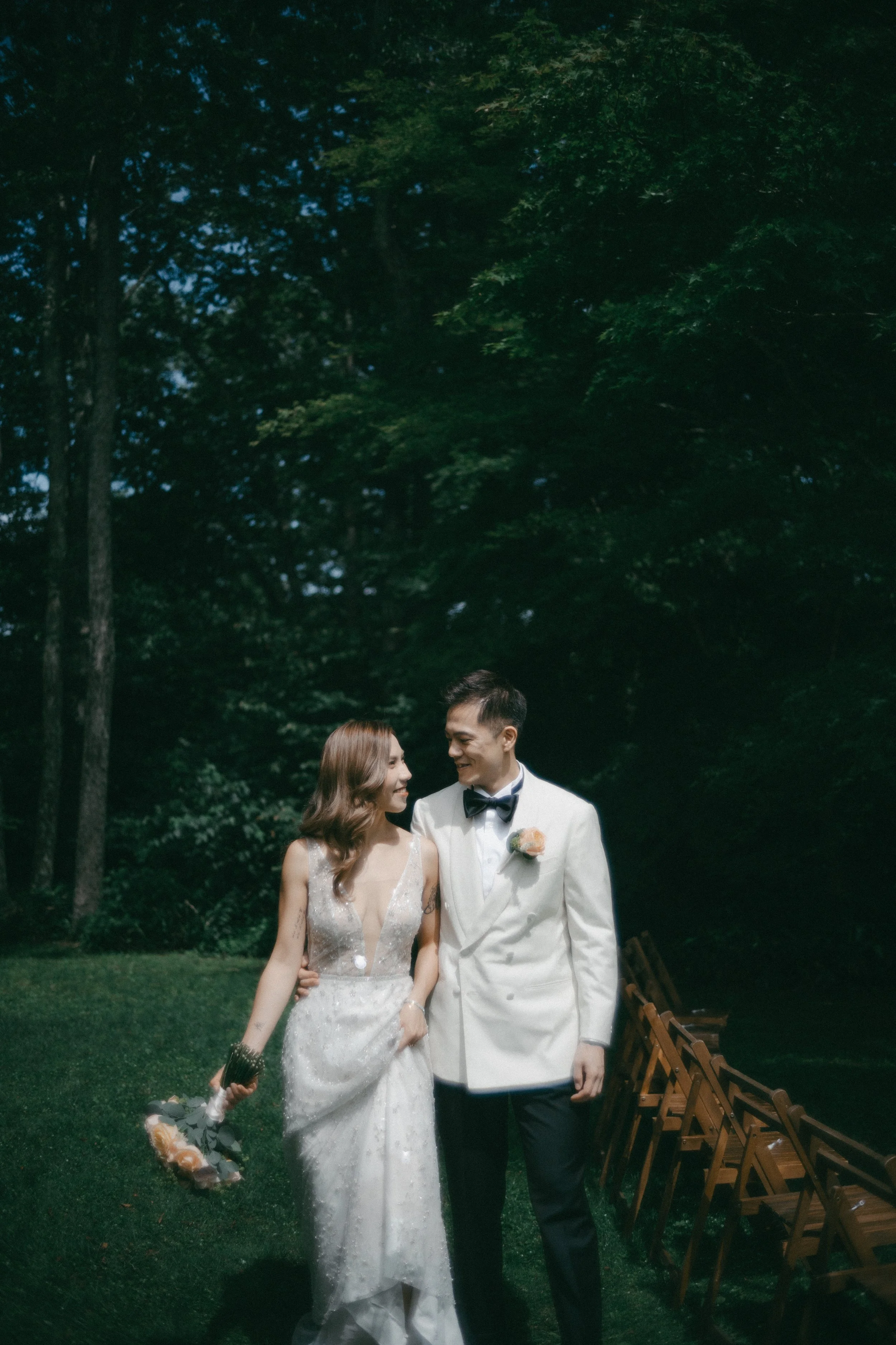 A bride and groom walking together outdoors, looking at each other and smiling, with trees in the background and chairs on the side.