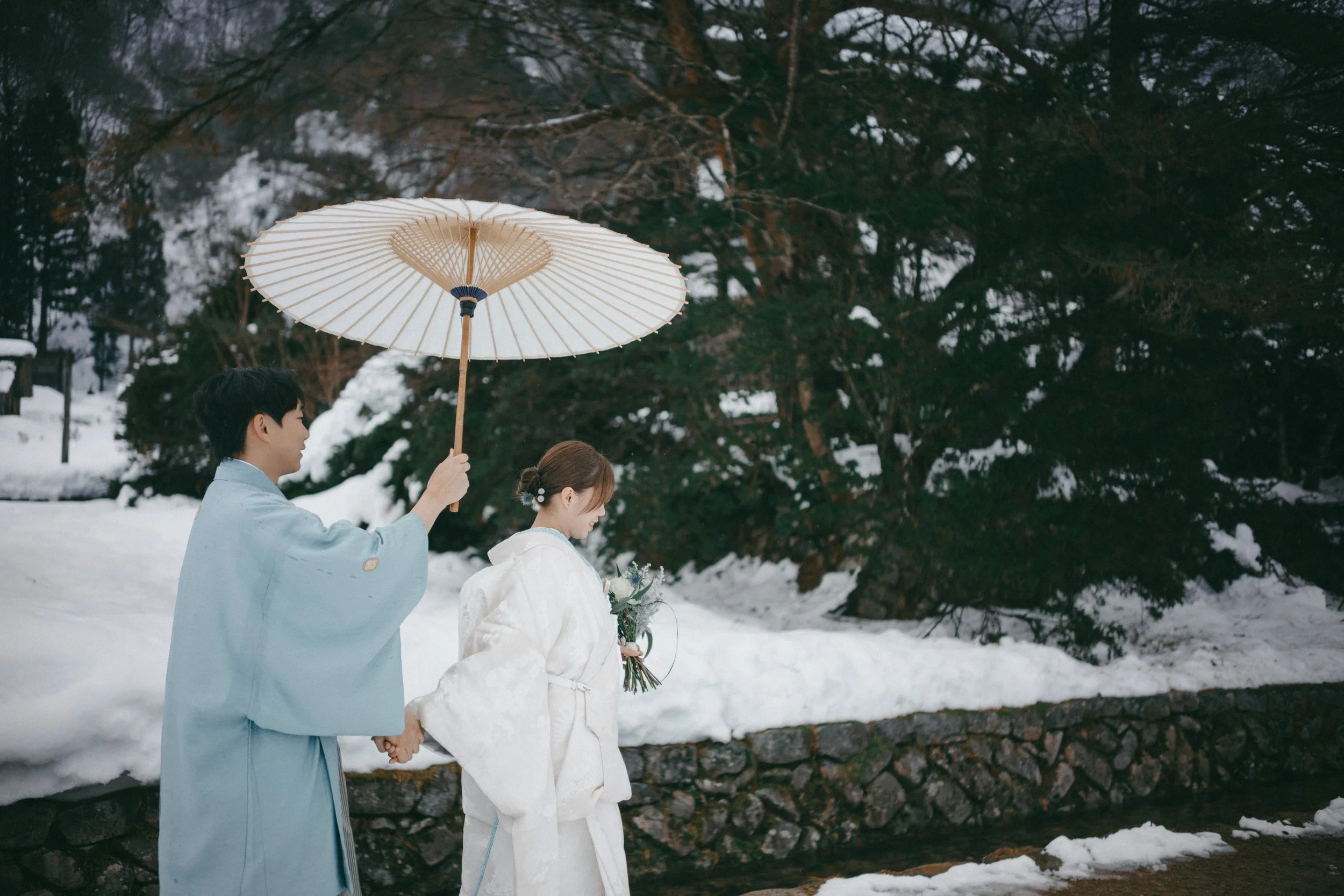 A couple dressed in traditional Japanese wedding attire walking outdoors in snow, with the groom holding a traditional paper parasol