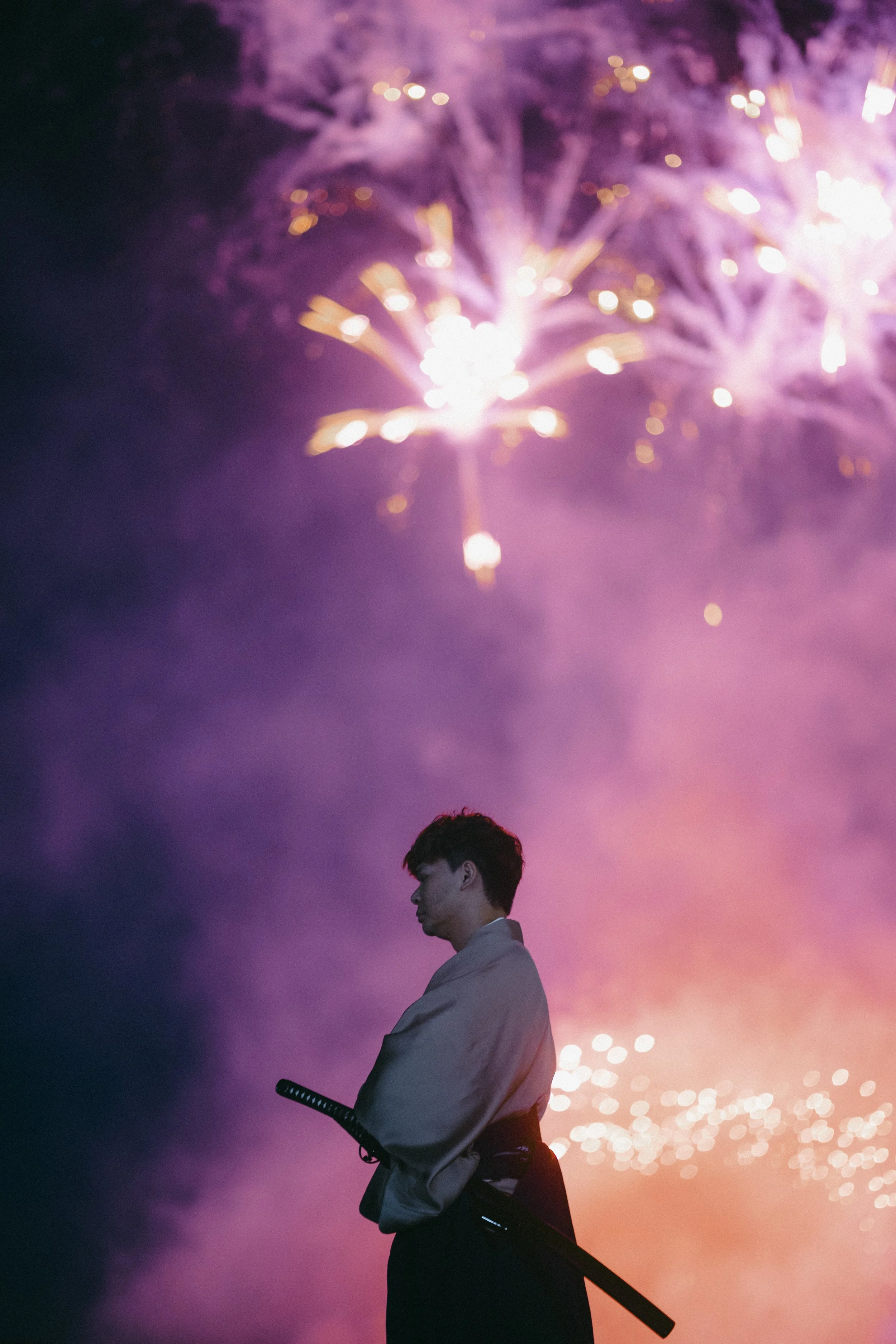 A person in traditional clothing holding a sword staring outdoors at fireworks lighting up the sky.