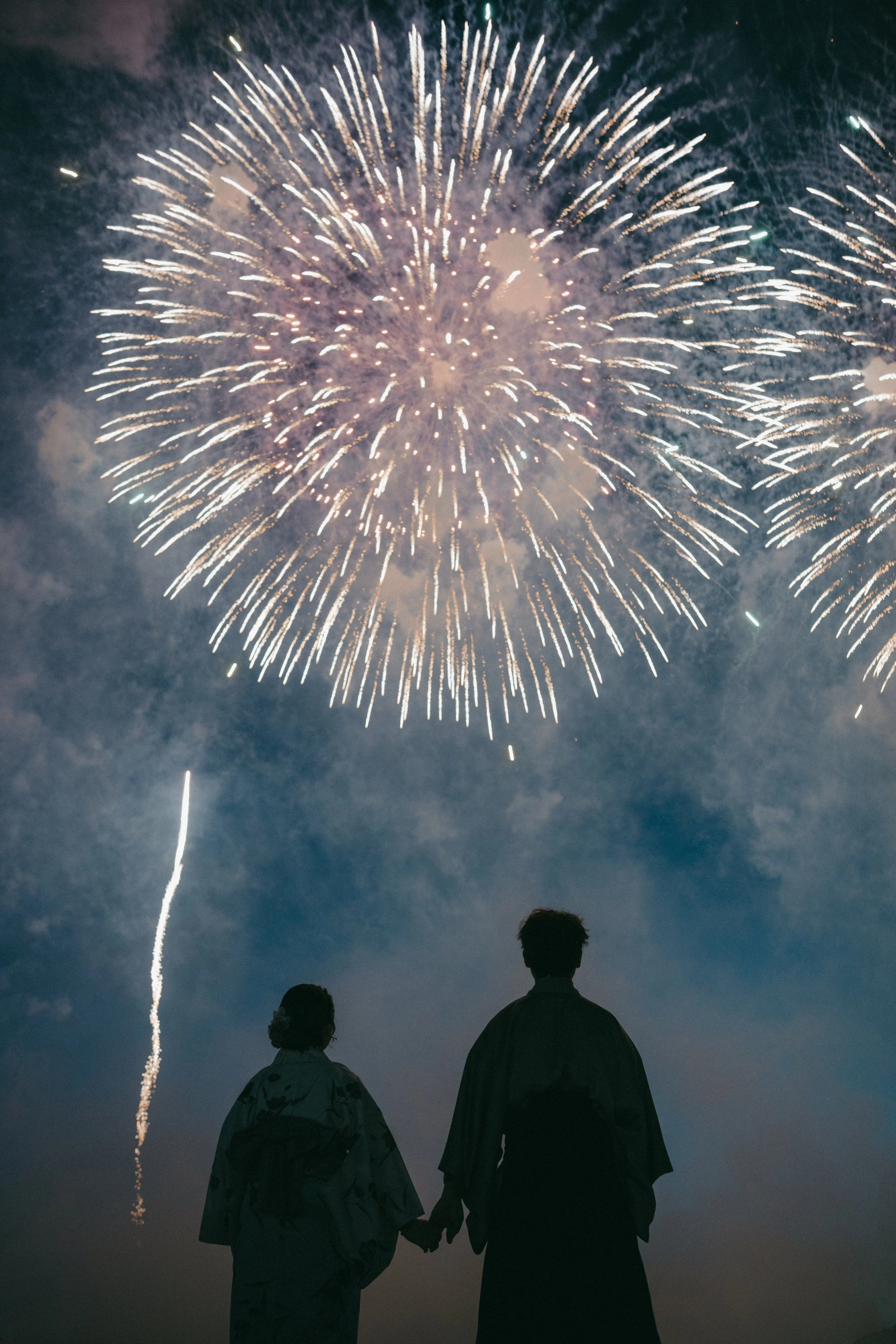 Silhouettes of a man and woman holding hands and watching fireworks in the night sky.