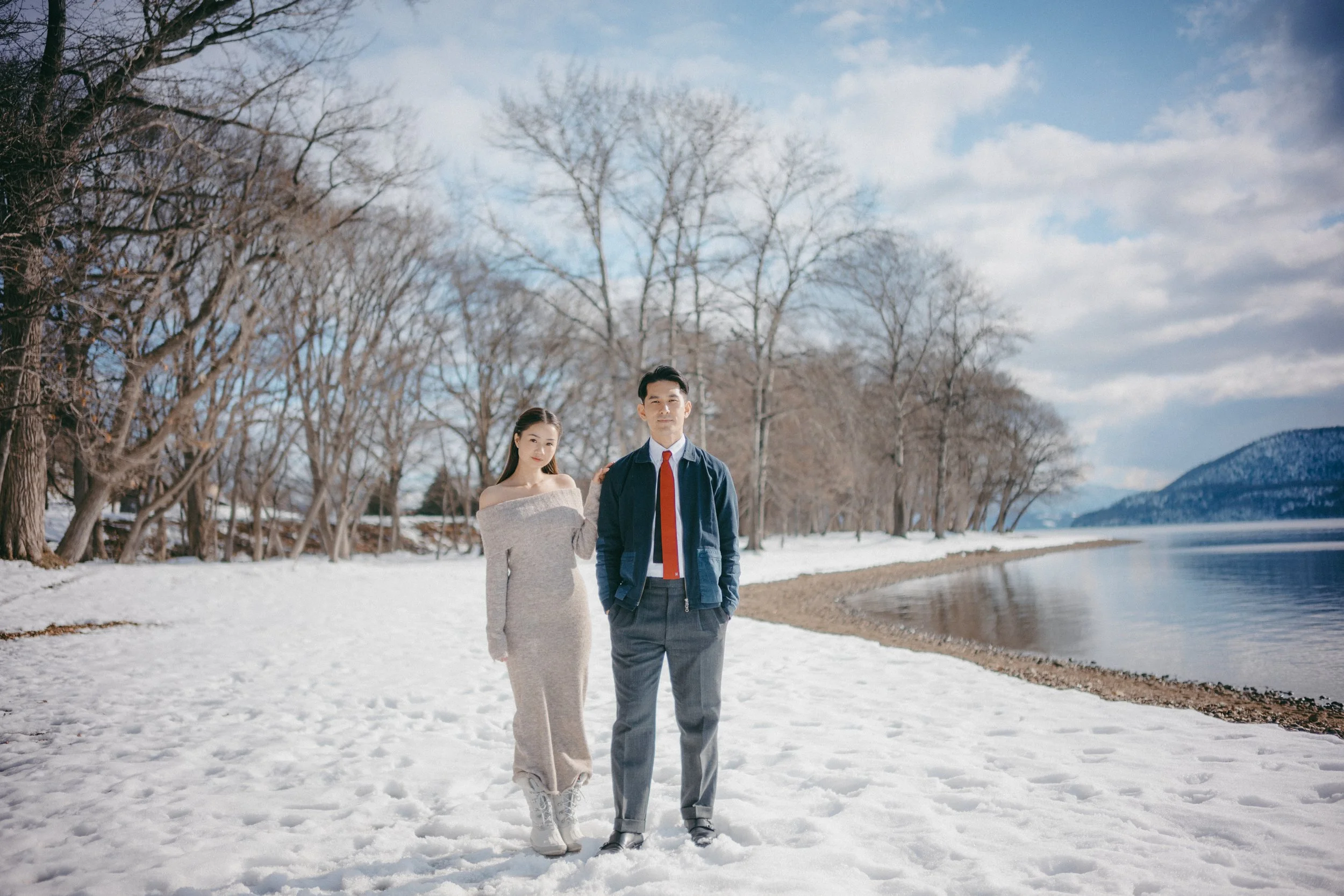 A man and woman standing on a snowy lakeside with bare trees and mountains in the background.