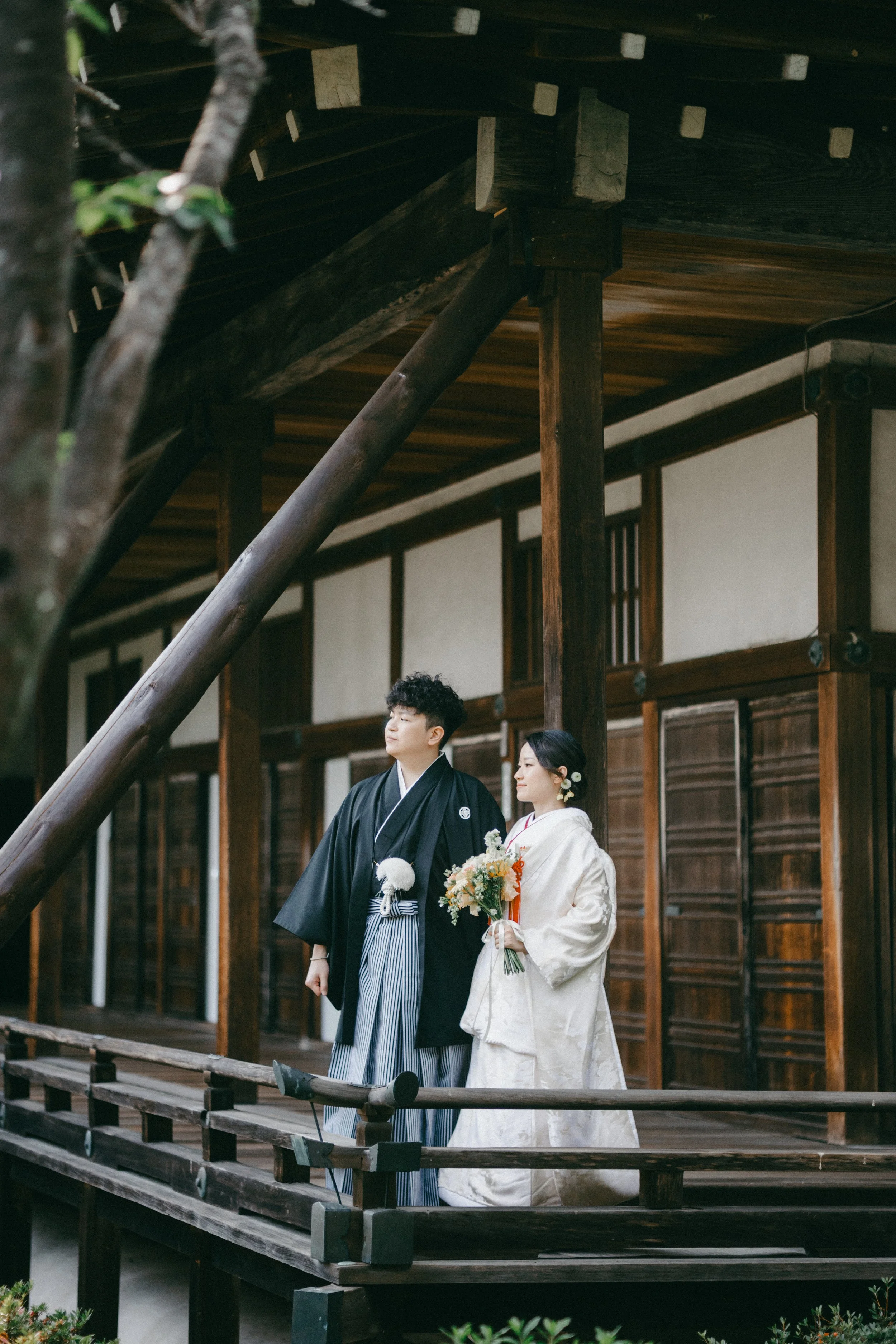 A couple dressed in traditional Japanese wedding attire standing on a wooden porch of a historic building, holding a bouquet of flowers.